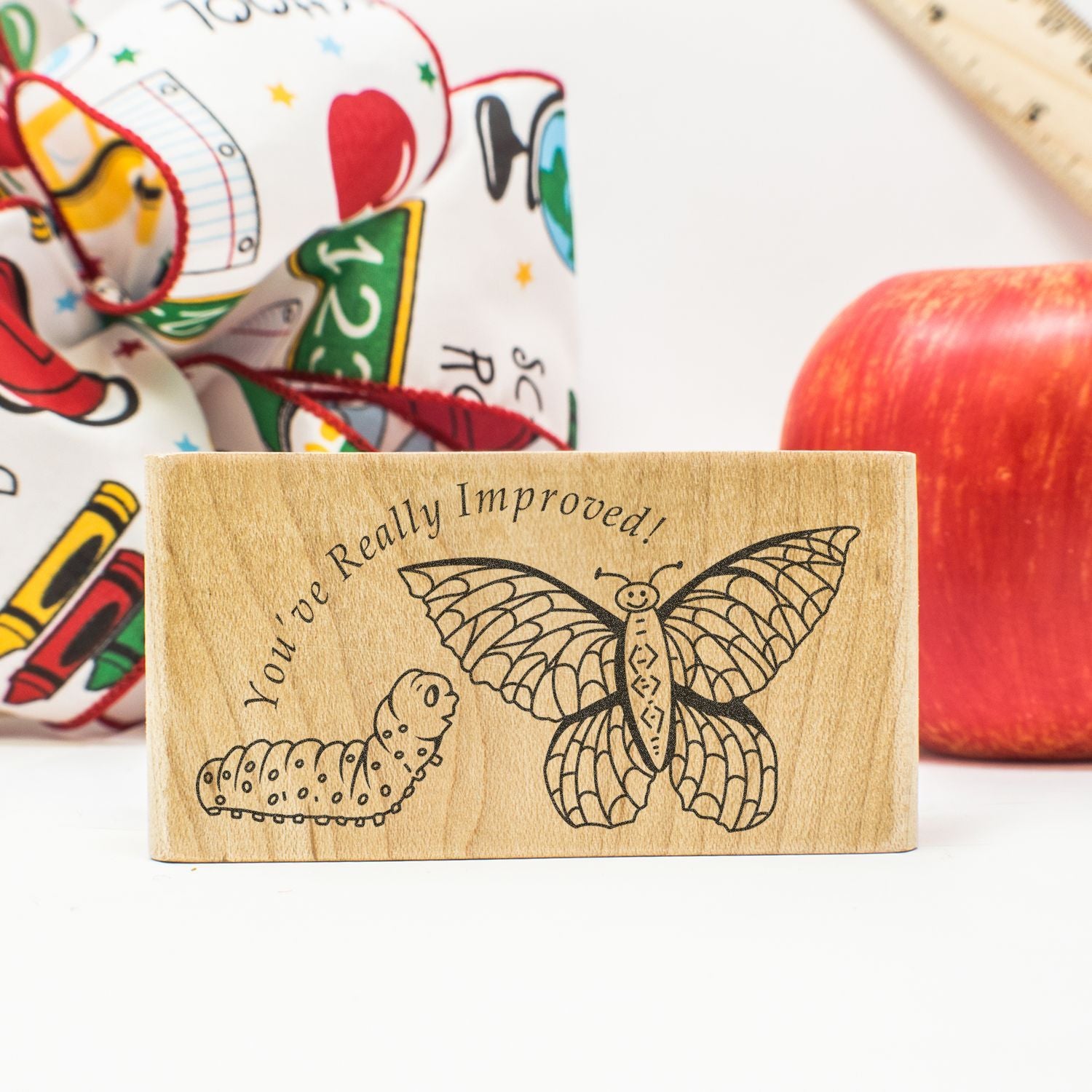Wooden butterfly and caterpillar rubber stamp with Youve Really Improved! text, school supplies and apple in background