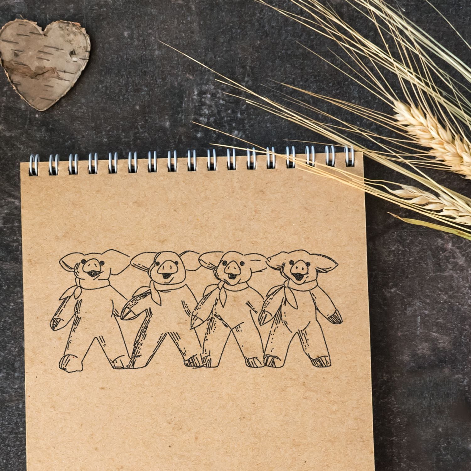 Kraft spiral notebook with cute pig teddy bear drawings, rustic heart decor, and wheat stalks on a dark background.