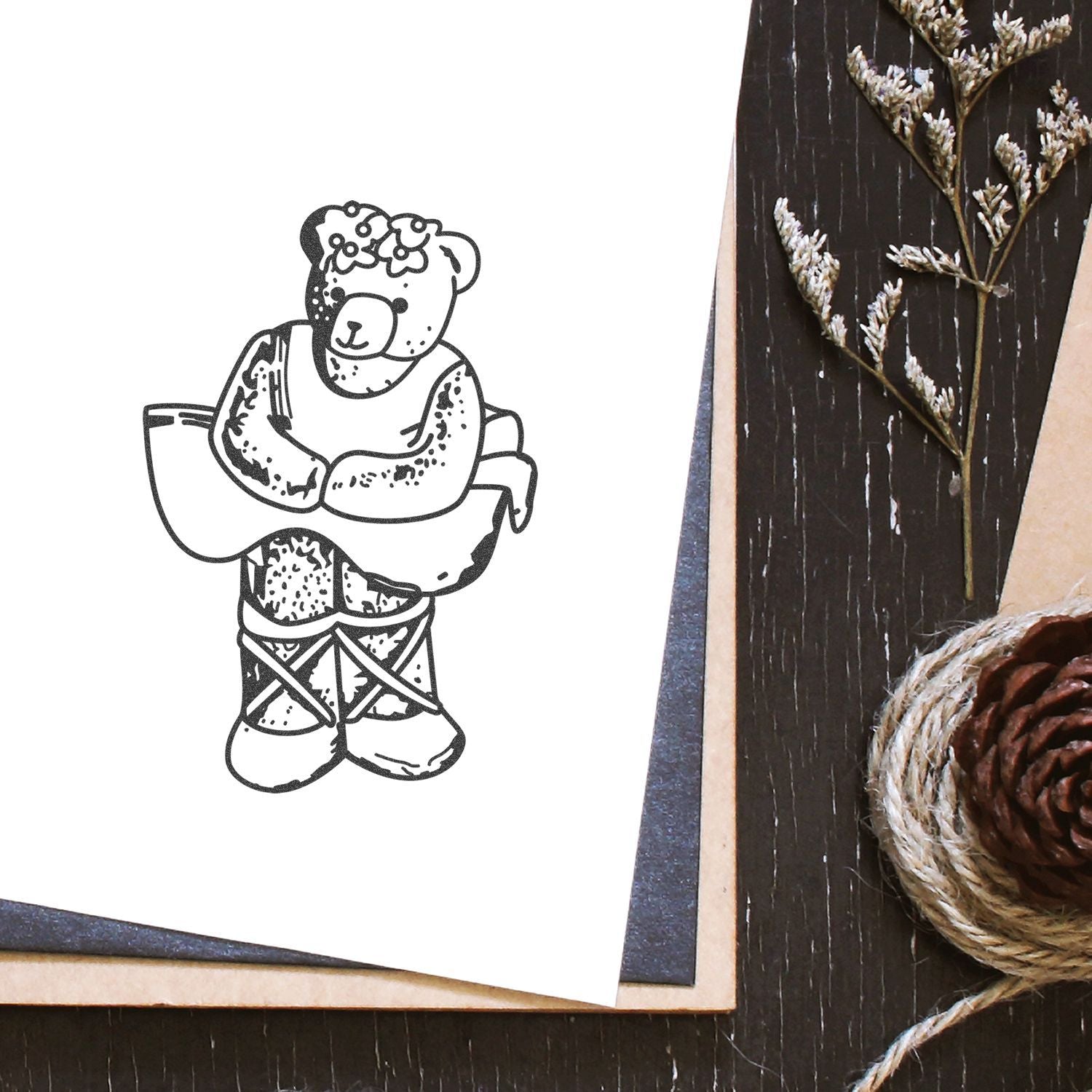 Black and white teddy bear illustration with floral crown on craft paper, styled with dried botanicals, pinecone, and twine on a rustic wooden table.