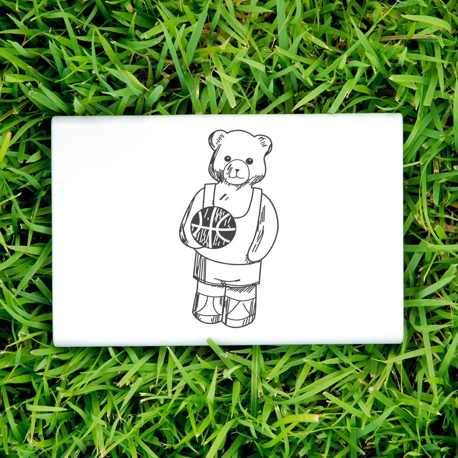 White laptop with cute teddy bear basketball design on green grass, sporty animal illustration.