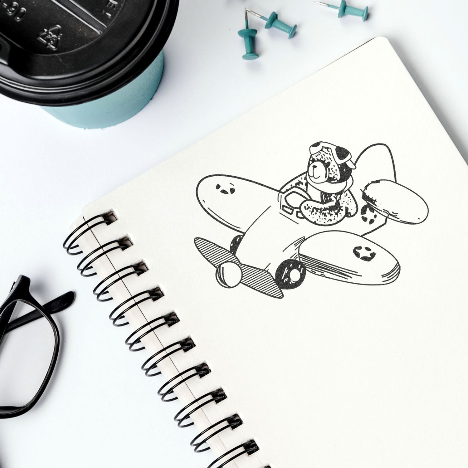 Spiral notebook with airplane and dog illustration, coffee cup, eyeglasses, and push pins on a clean white desk.