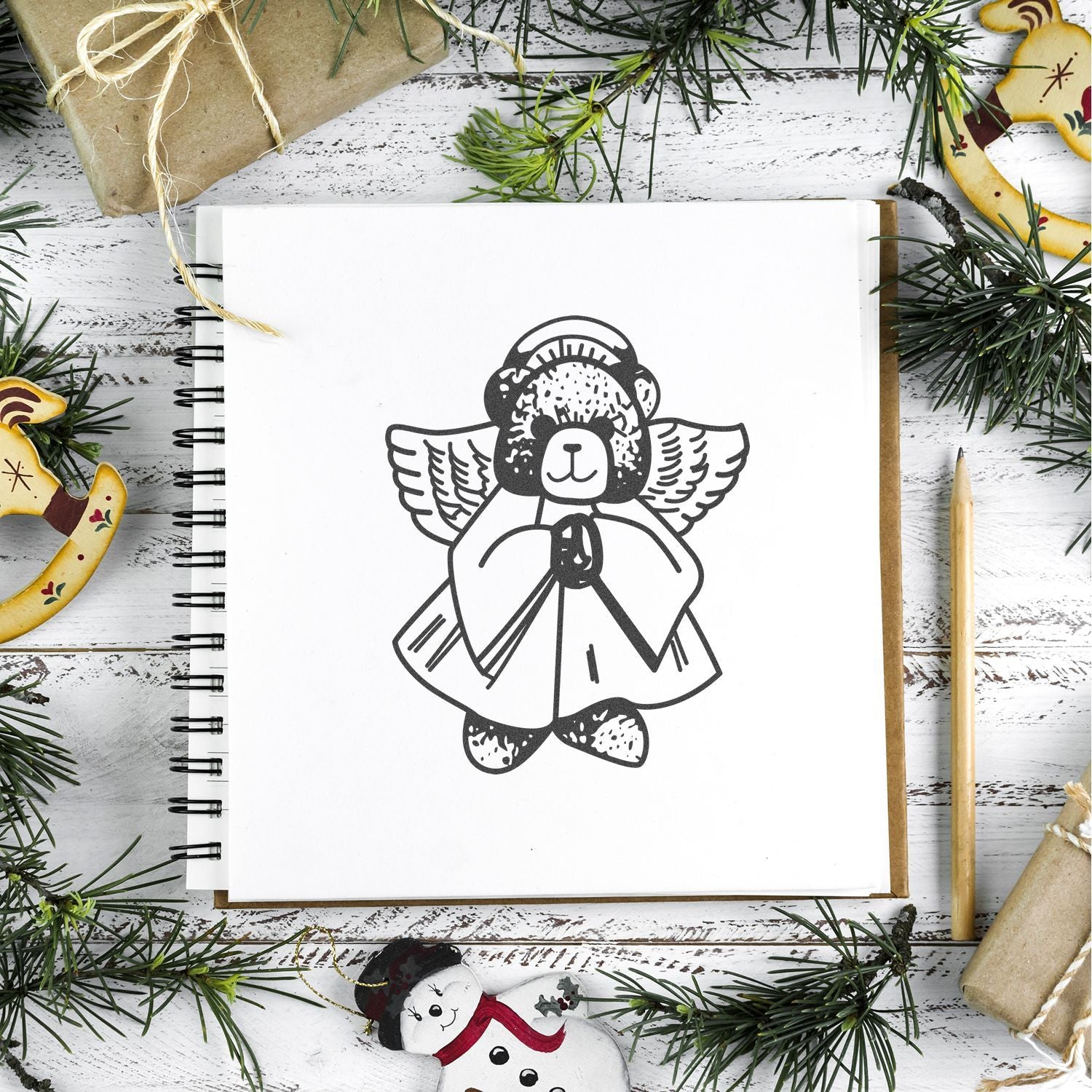 Holiday sketchbook with angel teddy bear drawing, surrounded by pine branches, wrapped gifts, and festive ornaments on a rustic white wooden background.