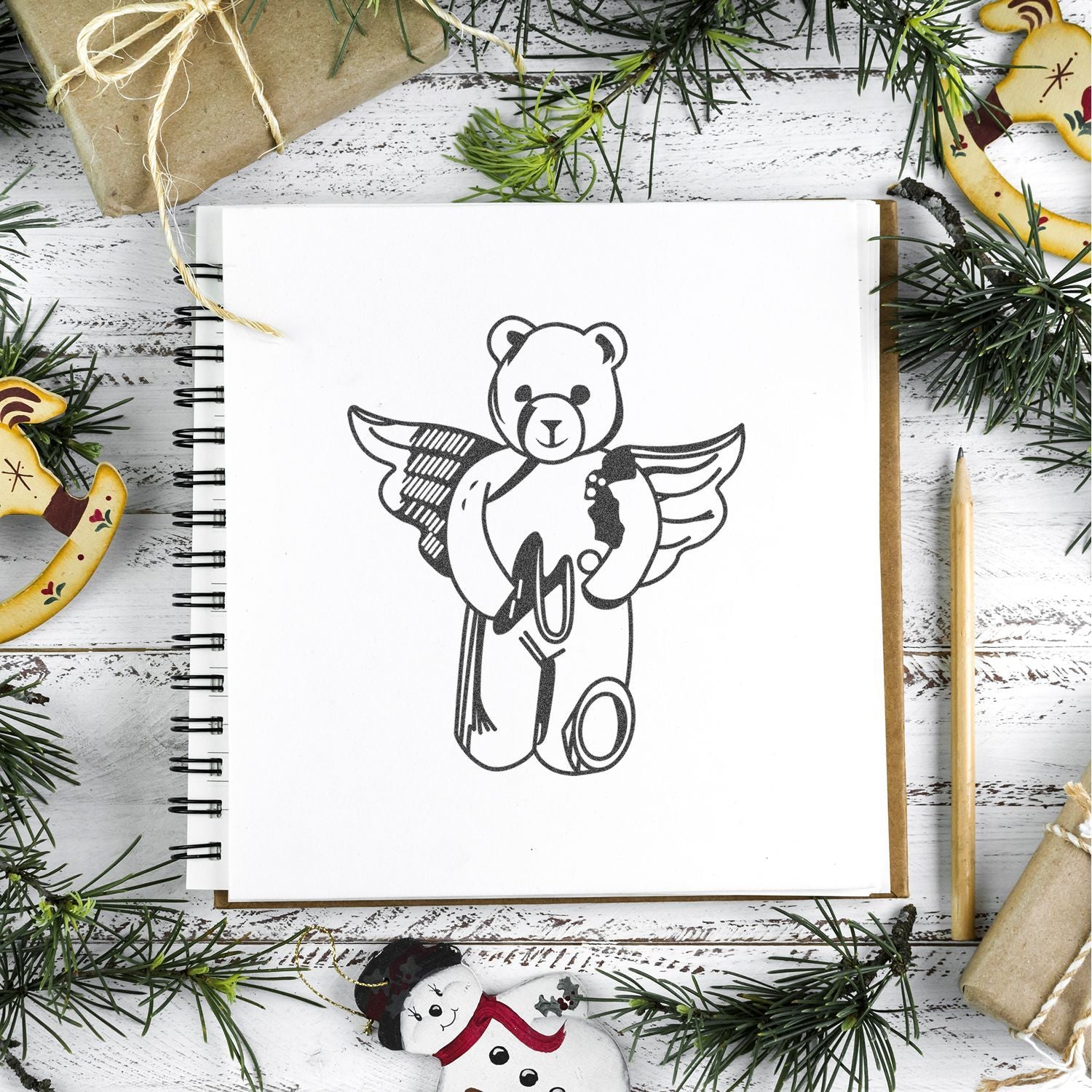 Holiday flat lay of a sketchbook with teddy bear angel drawing, surrounded by Christmas greenery, wrapped gifts, pencil, and ornaments on a rustic white wood background.
