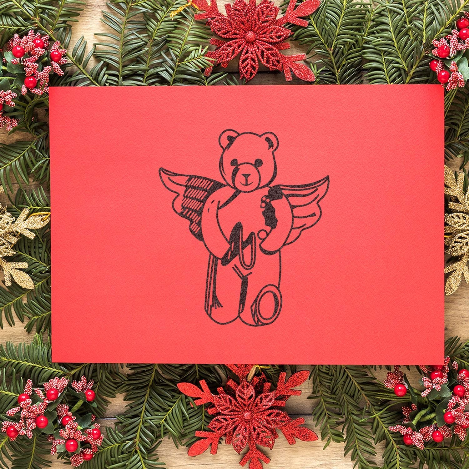 Red Christmas card with teddy bear angel design, surrounded by festive pine greenery and holiday decorations.