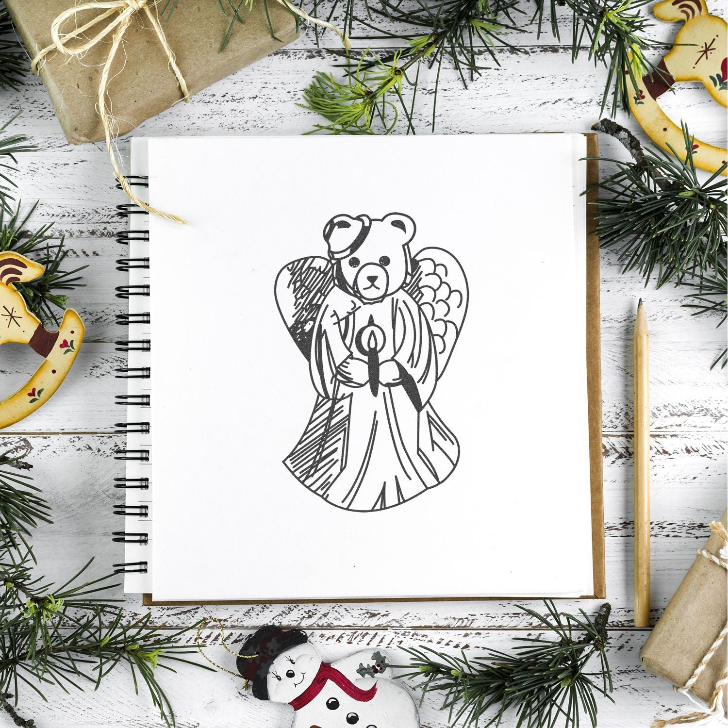 Hand-drawn bear angel sketch in a notebook surrounded by Christmas greenery, wrapped gifts, and holiday ornaments on a rustic white table.