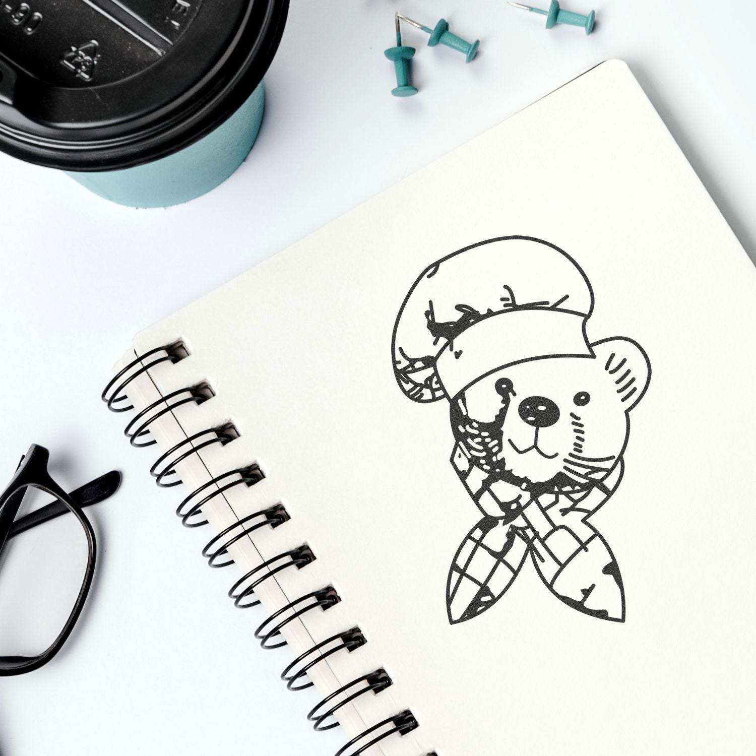 Spiral notebook with cute bear illustration, coffee cup, glasses, and push pins on a clean white desk flat lay.