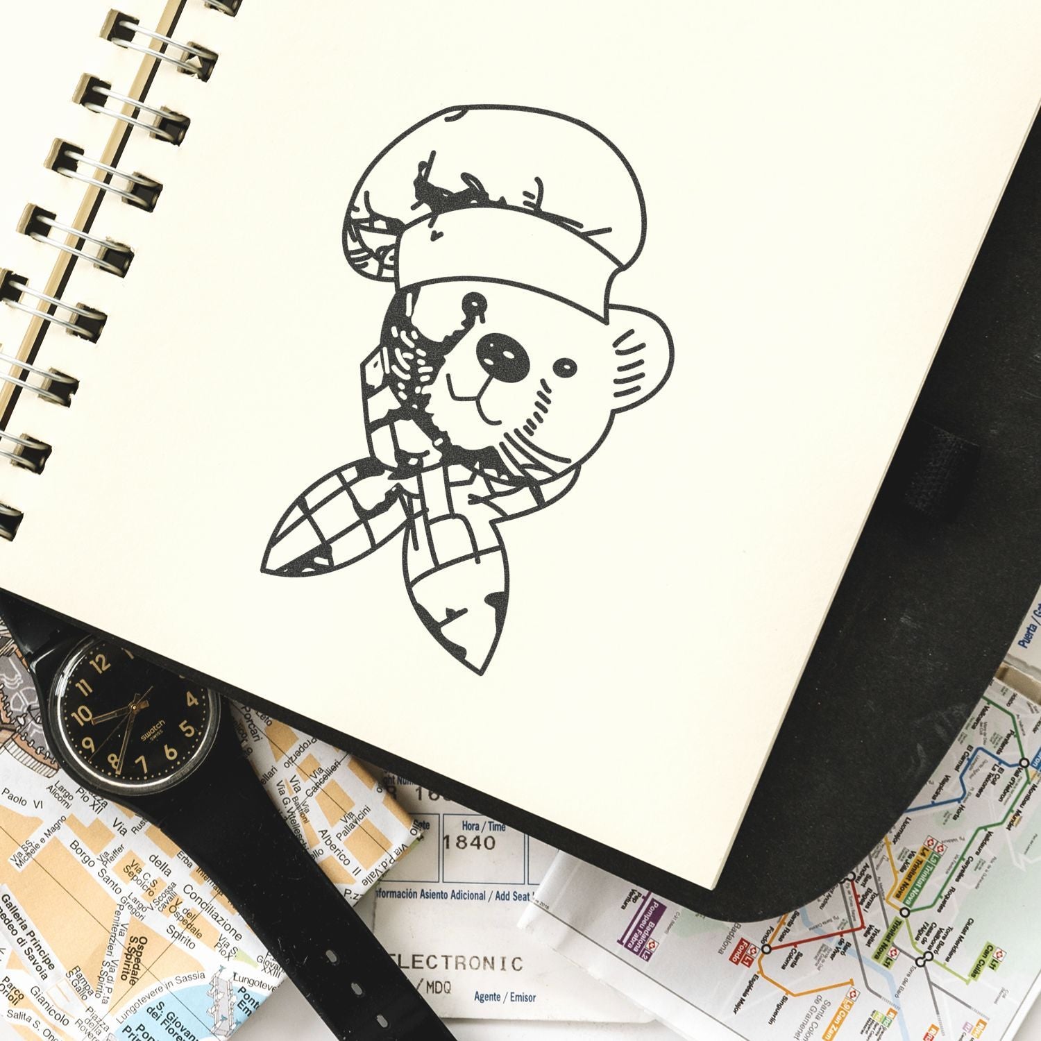 Black bear cartoon sketch in a spiral notebook with travel maps and a wristwatch, creative stationery flat lay.
