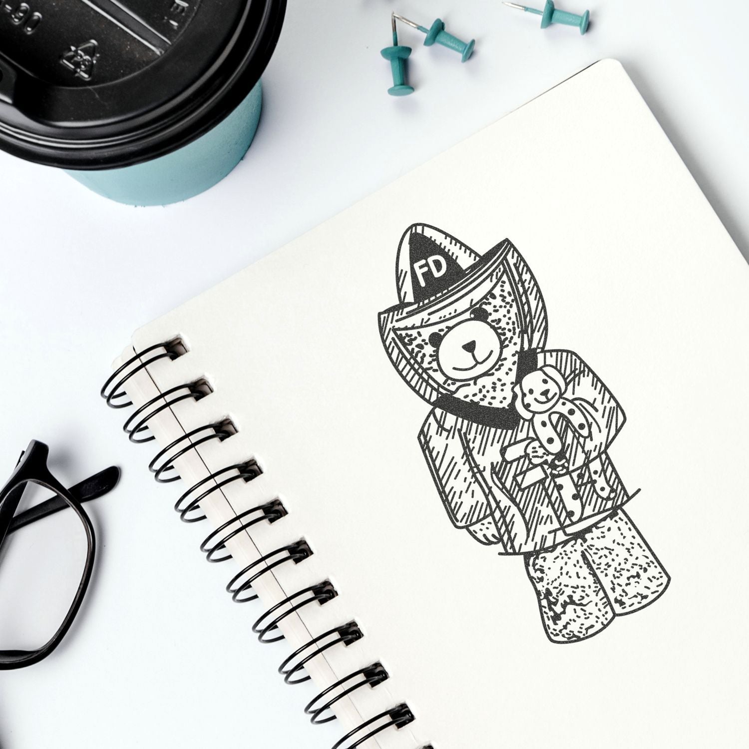Spiral notebook with cute firefighter teddy bear sketch, coffee cup, glasses, and push pins on a white desk.