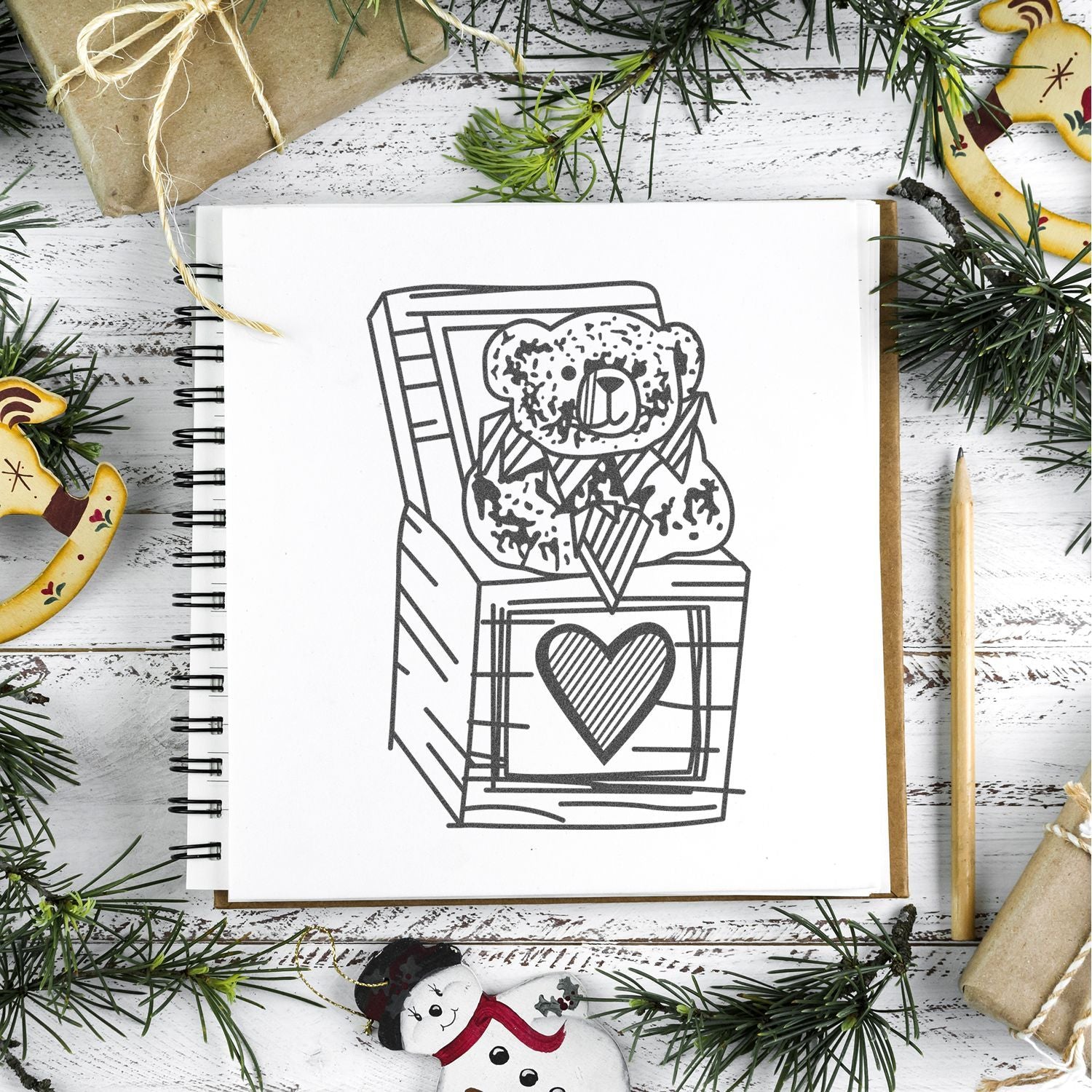Christmas-themed sketchbook with a bear in a gift box drawing, surrounded by pine branches, wrapped presents, and holiday decorations on a rustic white wooden background