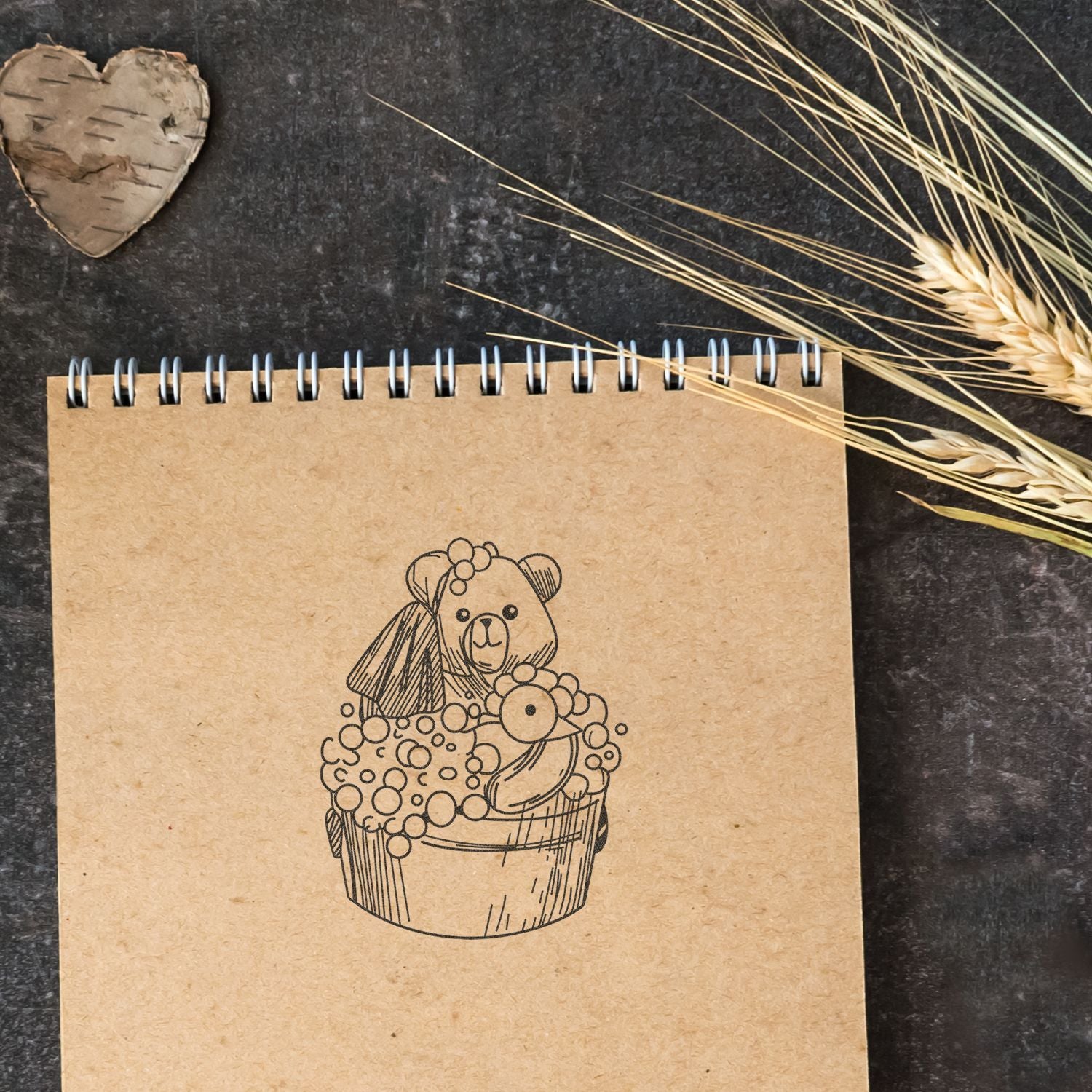 Kraft spiral notebook with cute teddy bear cupcake illustration on a rustic dark background with wheat and heart decor.