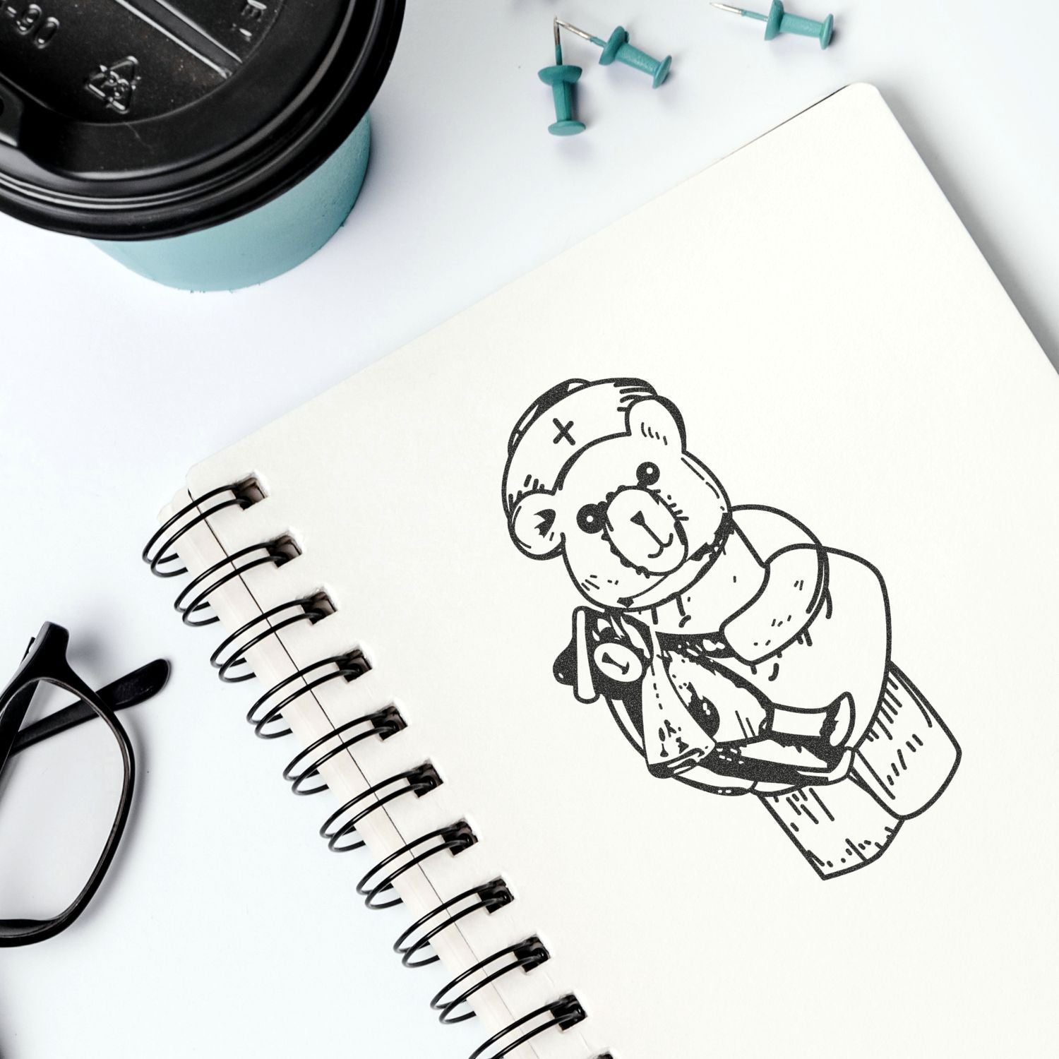 Flat lay of spiral notebook with cute bear sketch, coffee cup, glasses, and push pins on a white desk.