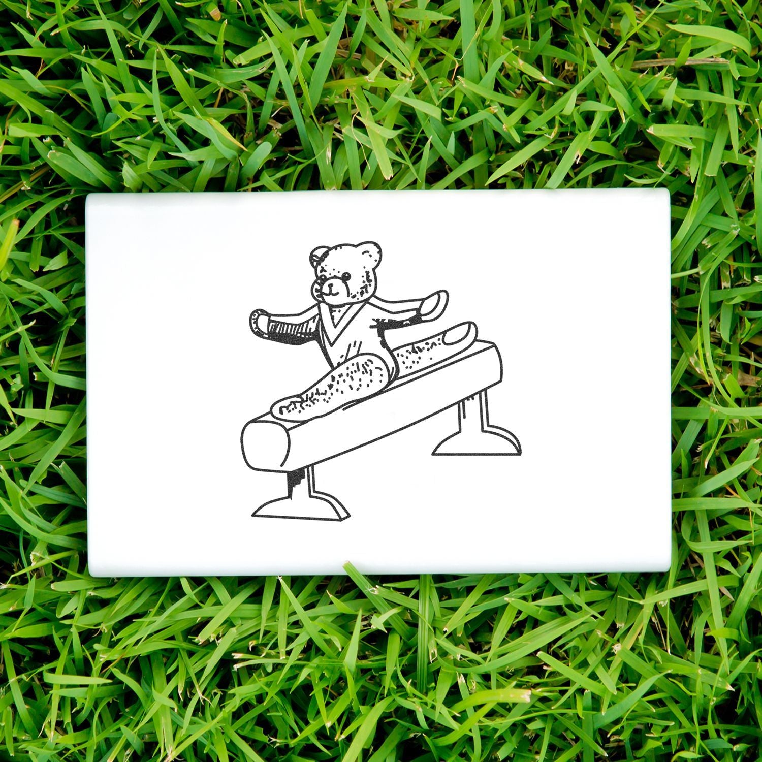 White ceramic tile with teddy bear gymnast line drawing on pommel horse in green grass.