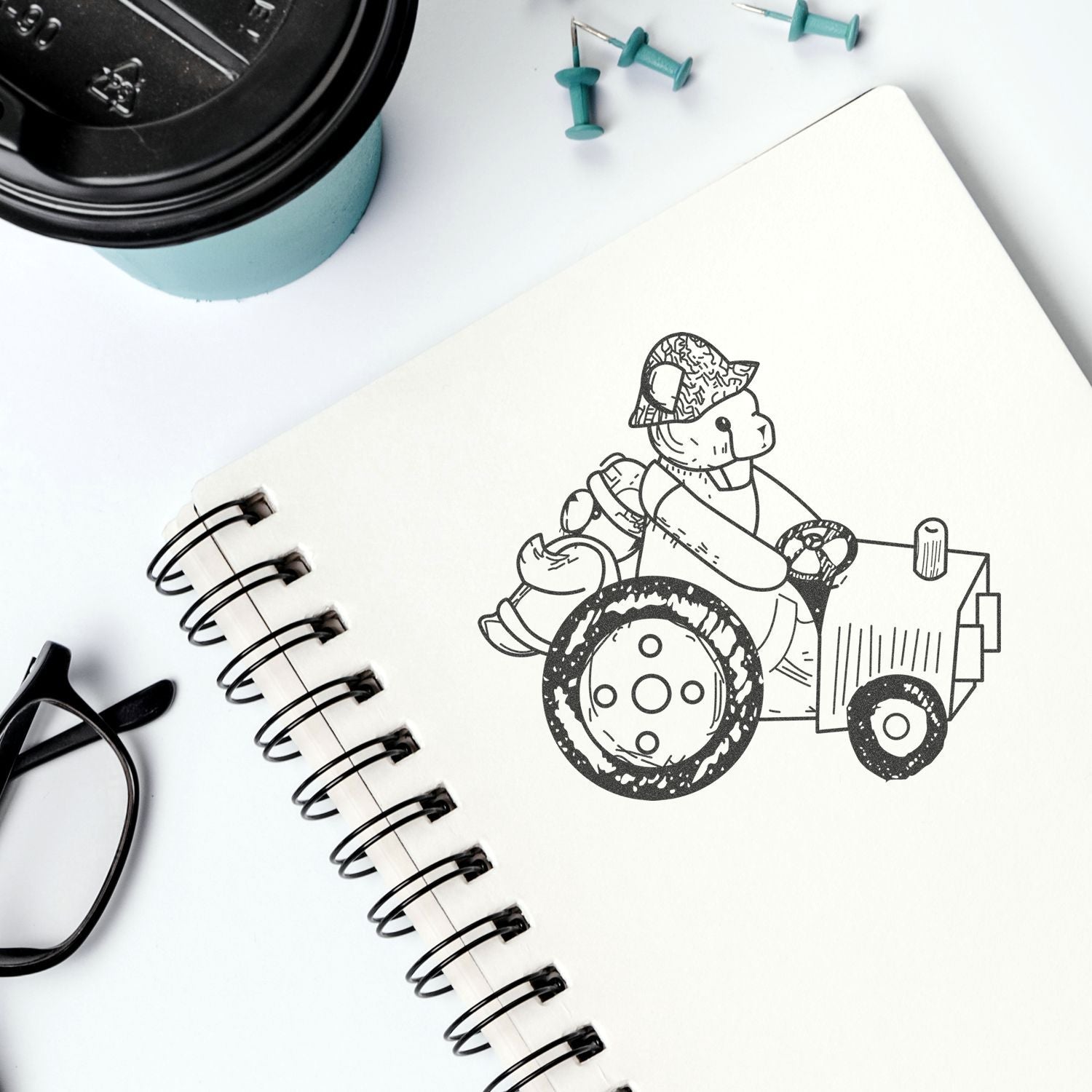 Sketchbook with cute bear driving a tractor drawing, coffee cup, glasses, and office supplies on a white desk.