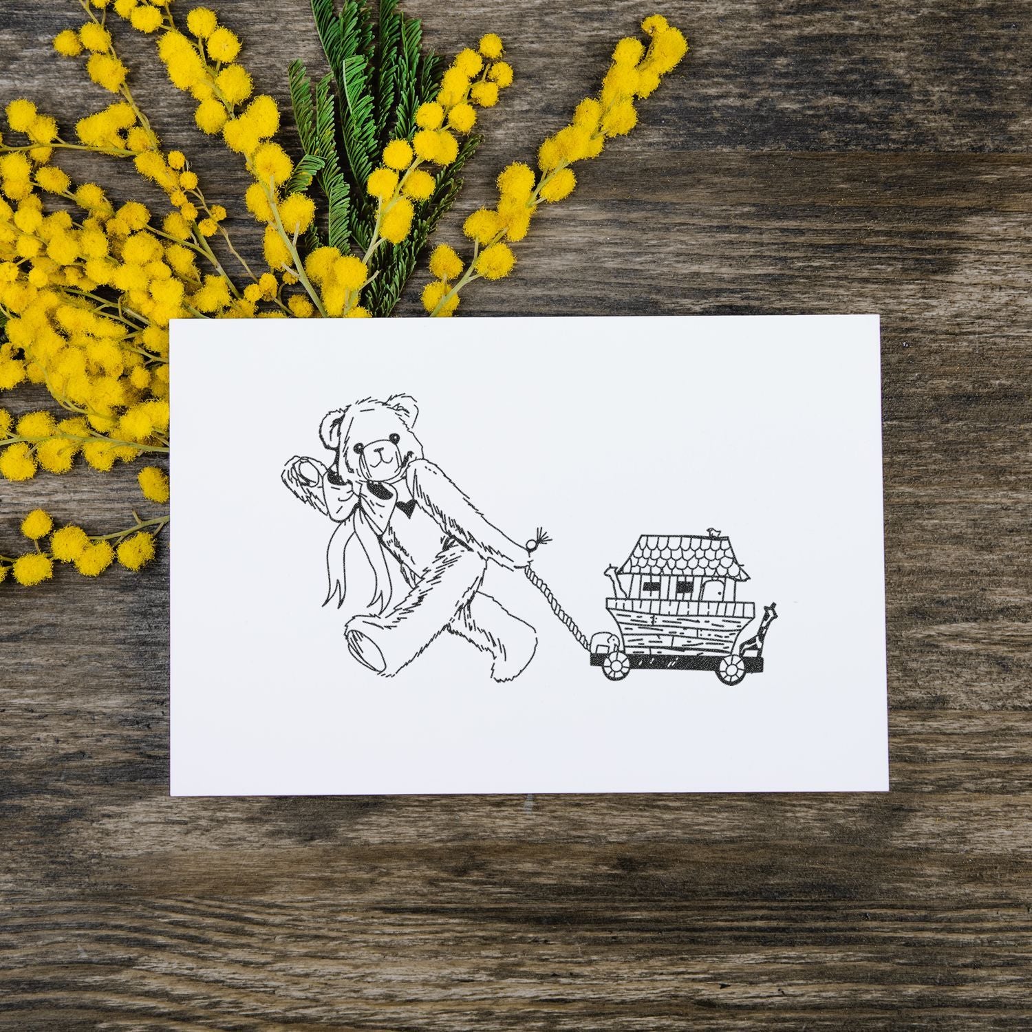 Teddy bear line art card with Noahs ark wagon, surrounded by yellow mimosa flowers on a rustic wooden background.