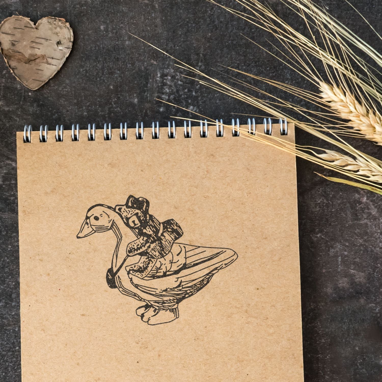 Kraft spiral notebook with a duck illustration on a rustic dark background, styled with dried wheat and a wooden heart decor.