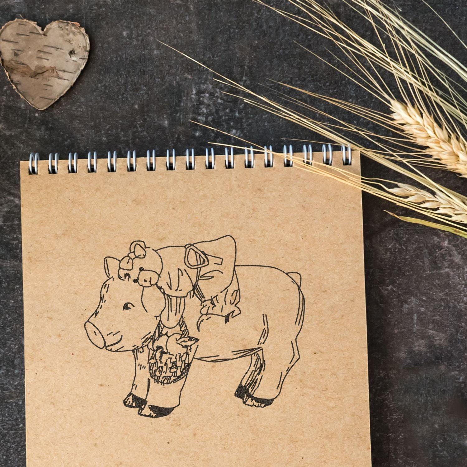 Spiral notebook with cute pig sketch on kraft paper, styled with wheat stalks and rustic heart decor on a dark background.