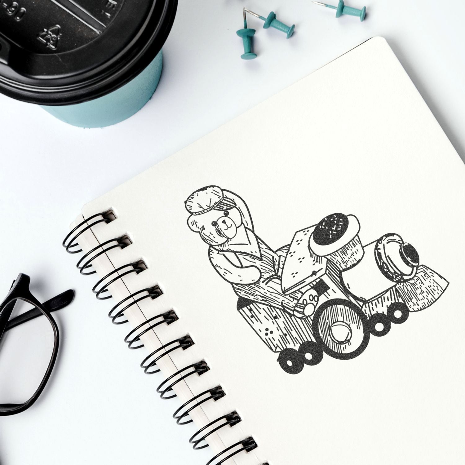 Minimal flat lay of a spiral notebook with a cute bear illustration, coffee cup, glasses, and stationery on a white desk background.