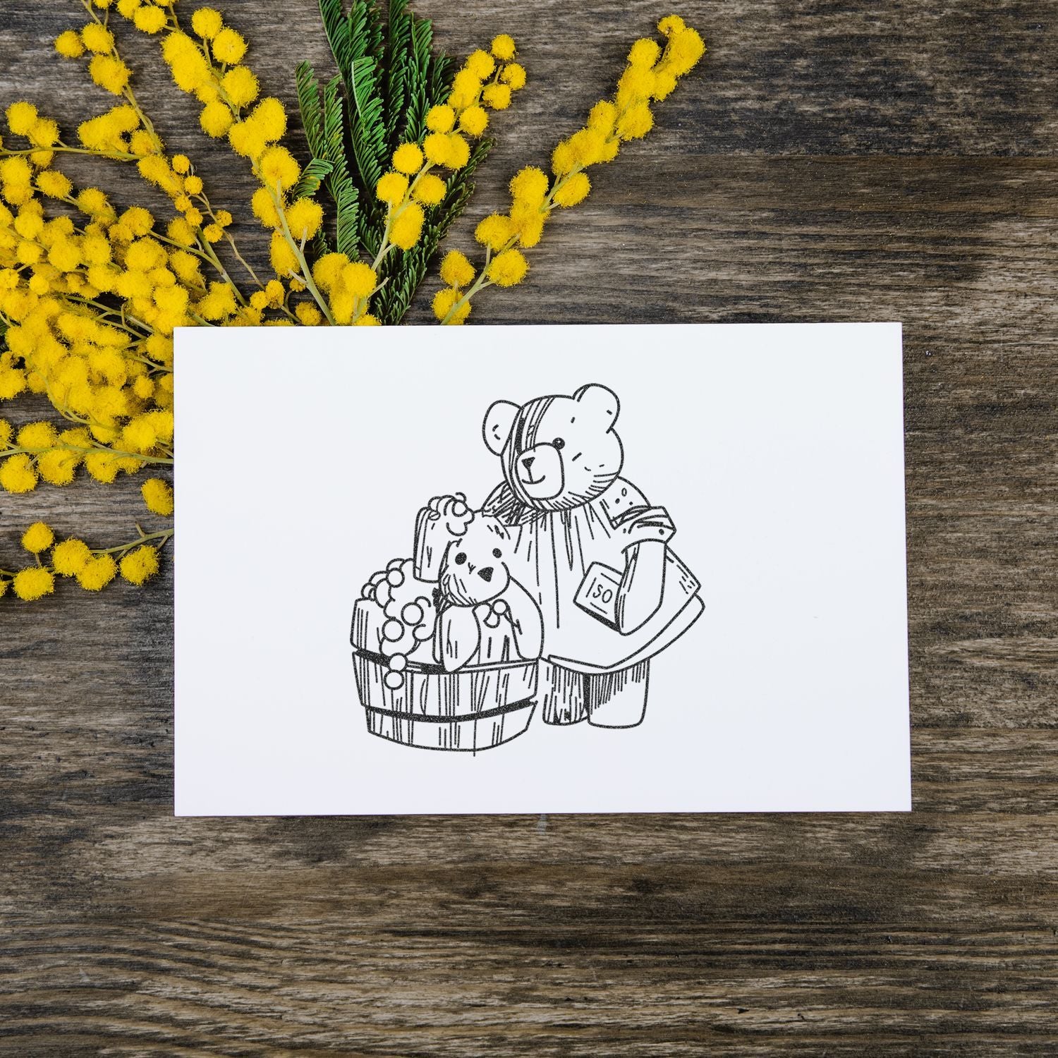 Hand-drawn teddy bear greeting card with yellow mimosa flowers on rustic wooden background.