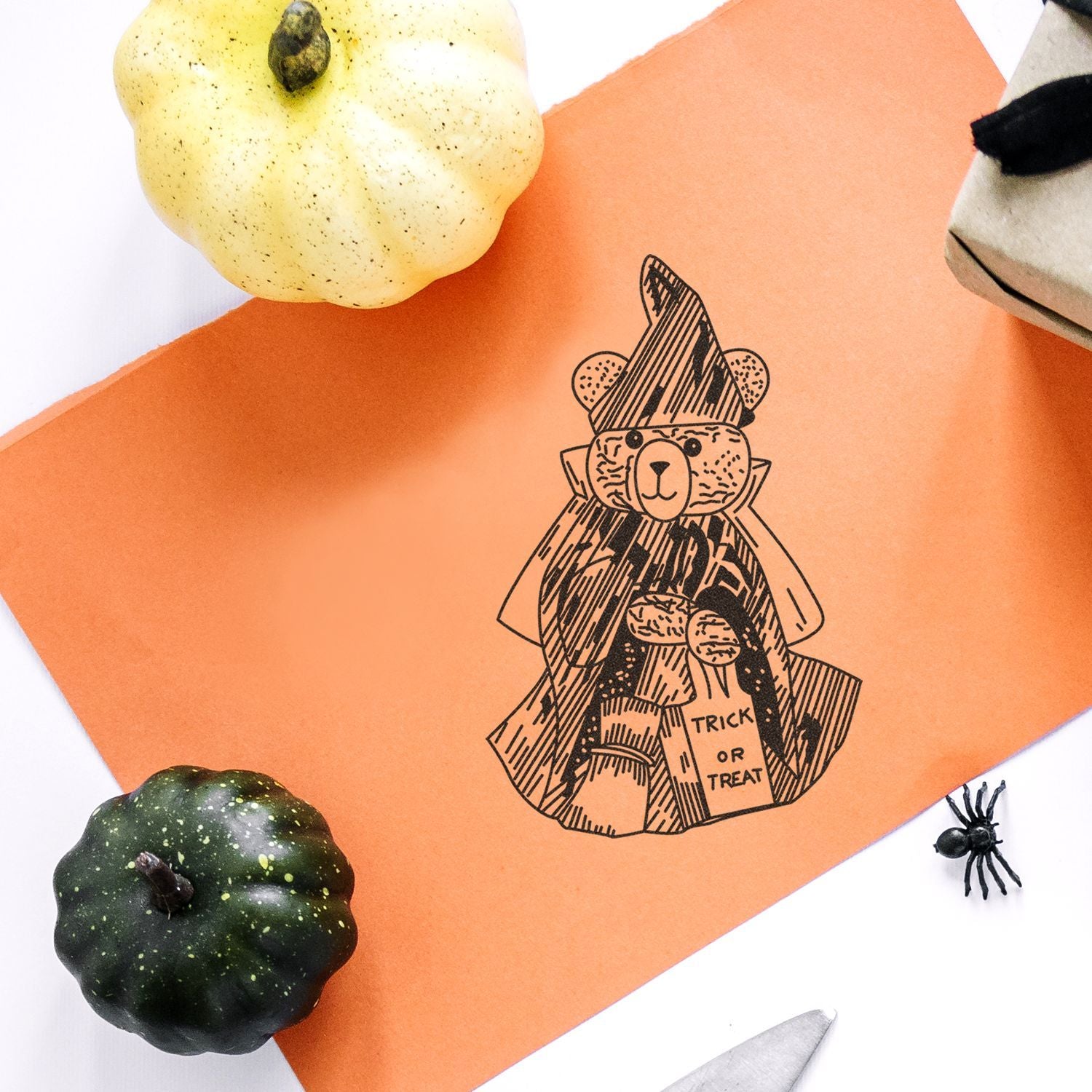 Halloween craft flat lay with orange paper, trick-or-treat bear illustration, decorative pumpkins, and a small spider.