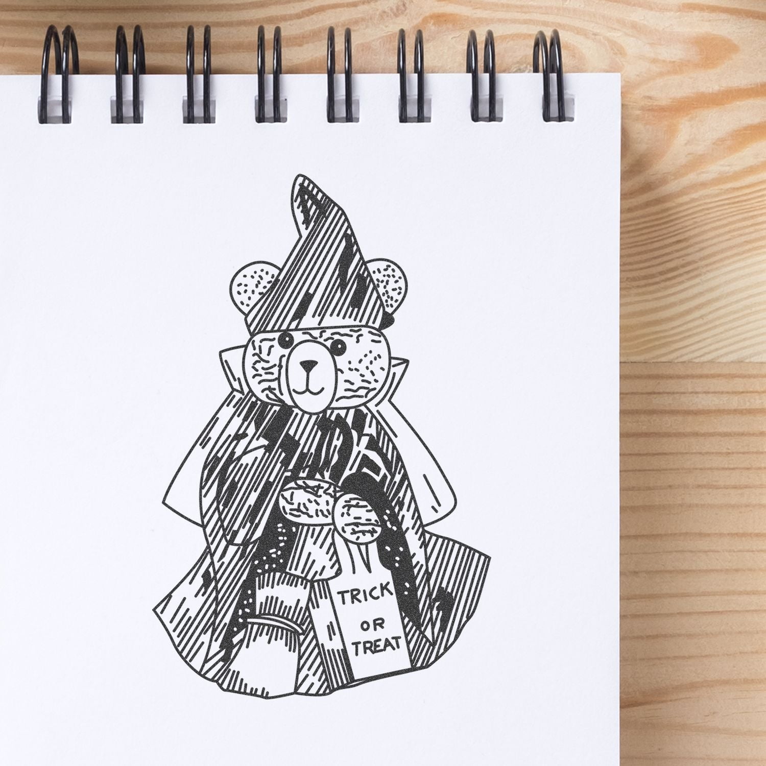 Black-and-white teddy bear Halloween costume sketch in a spiral notebook with trick or treat bag on wooden desk.