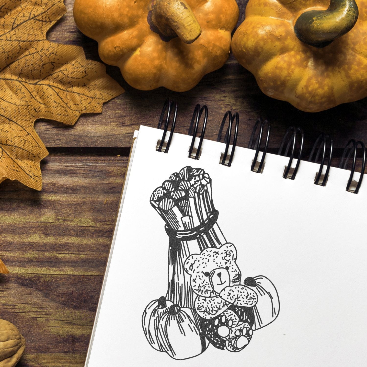 Autumn flat lay with decorative pumpkins, fall leaves, and a spiral notebook featuring a hand-drawn teddy bear pumpkin sketch on a rustic wooden background.