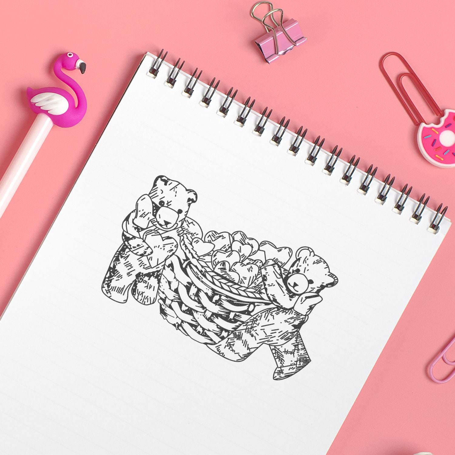 Spiral notebook with teddy bear drawing on pink desk, surrounded by flamingo pen, binder clips, and donut paperclips, cute stationery flat lay.