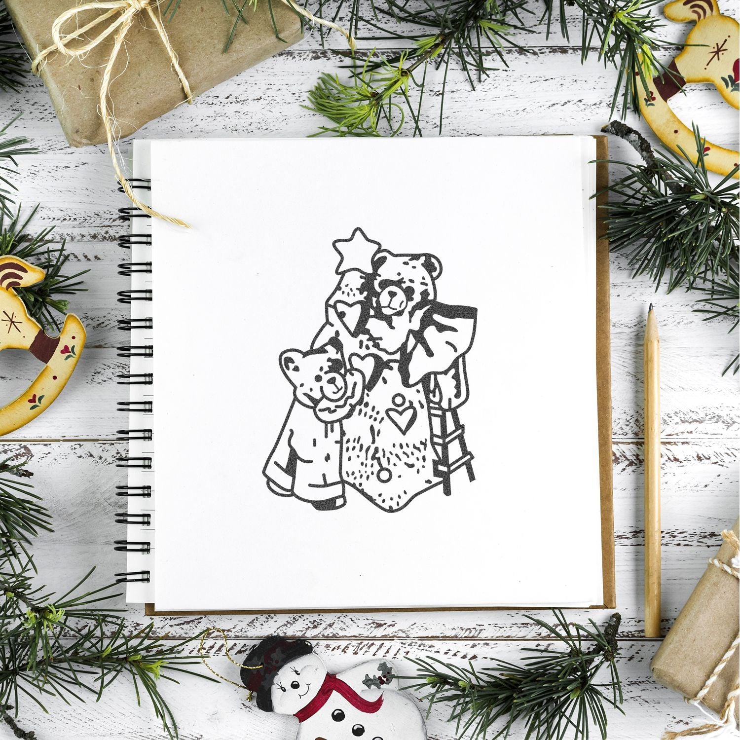Festive Christmas flat lay with a sketchbook featuring a teddy bear illustration, surrounded by pine branches, wrapped gifts, and holiday decorations.