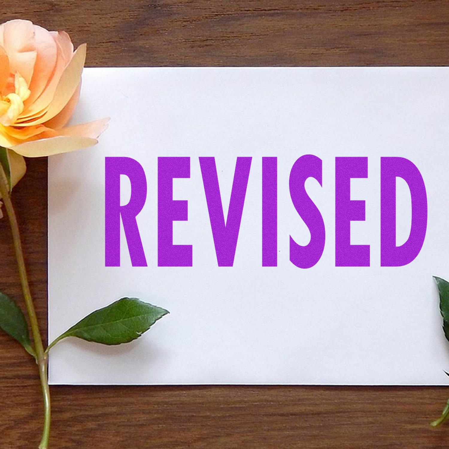 Purple REVISED text on a white card with flowers and green leaves on a wooden table.