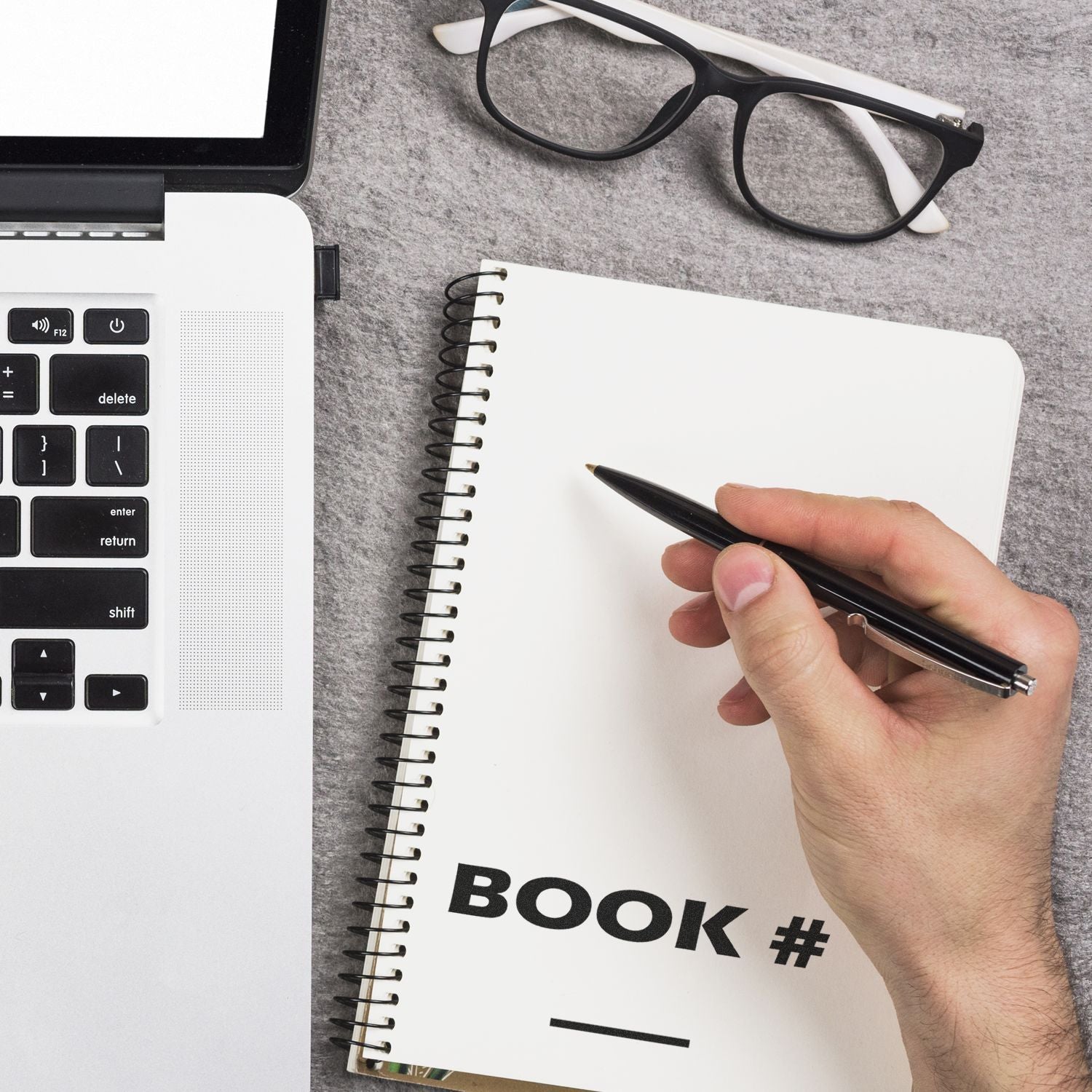 Hand writing in a notebook with a pen next to a laptop keyboard and glasses, illustrating productive desk note-taking for Book #.