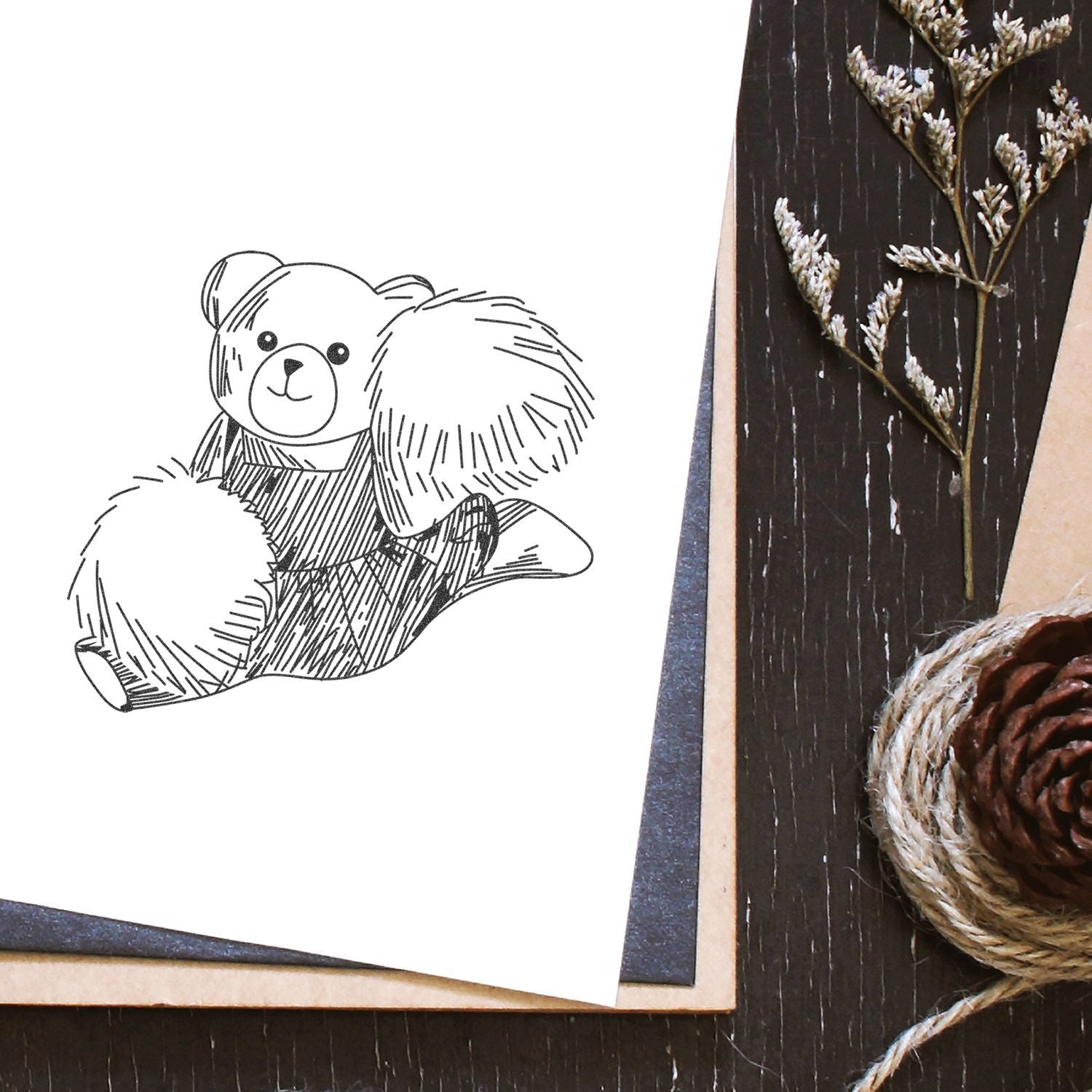 Teddy bear line drawing on white paper with rustic craft decor, pinecone, twine, and dried botanical branch on a dark wood background