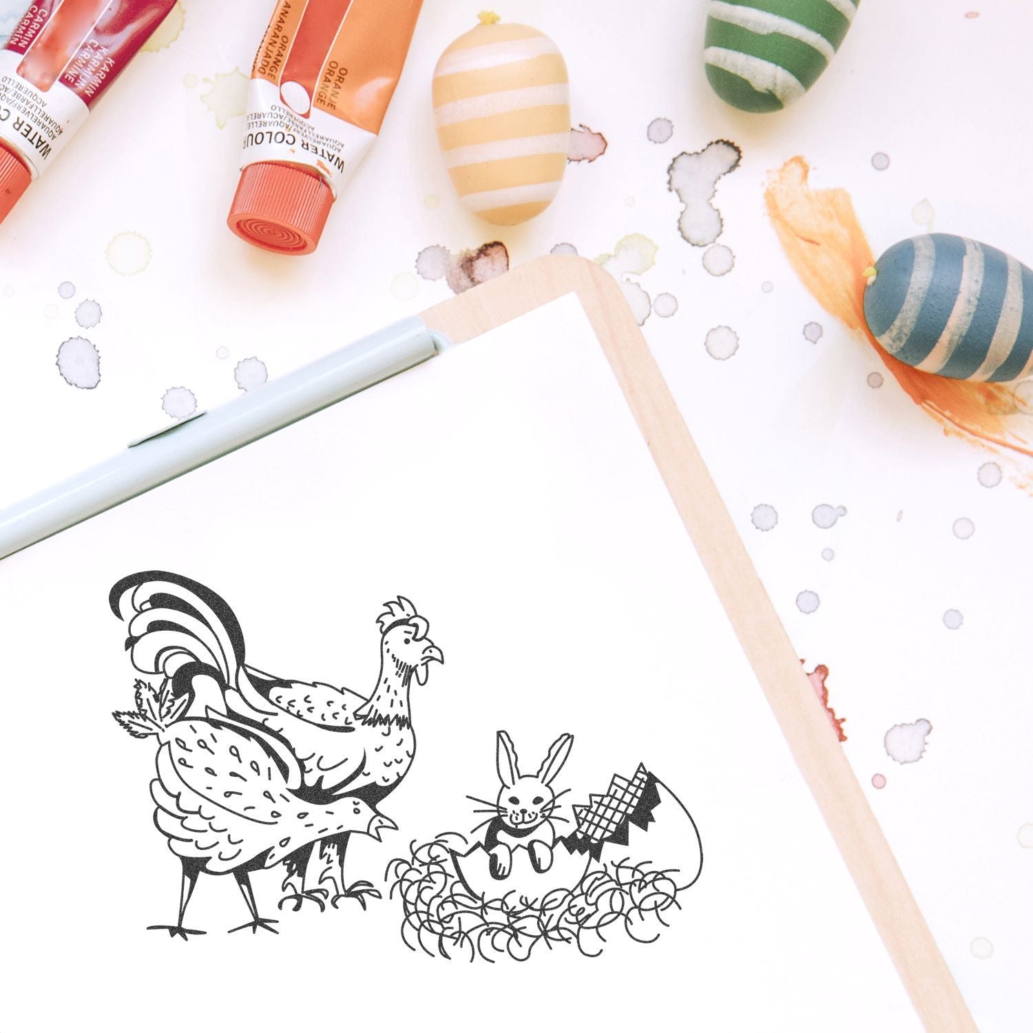 Watercolor painting supplies with tubes, decorated Easter eggs, and a sketchbook featuring a chicken and rabbit illustration.