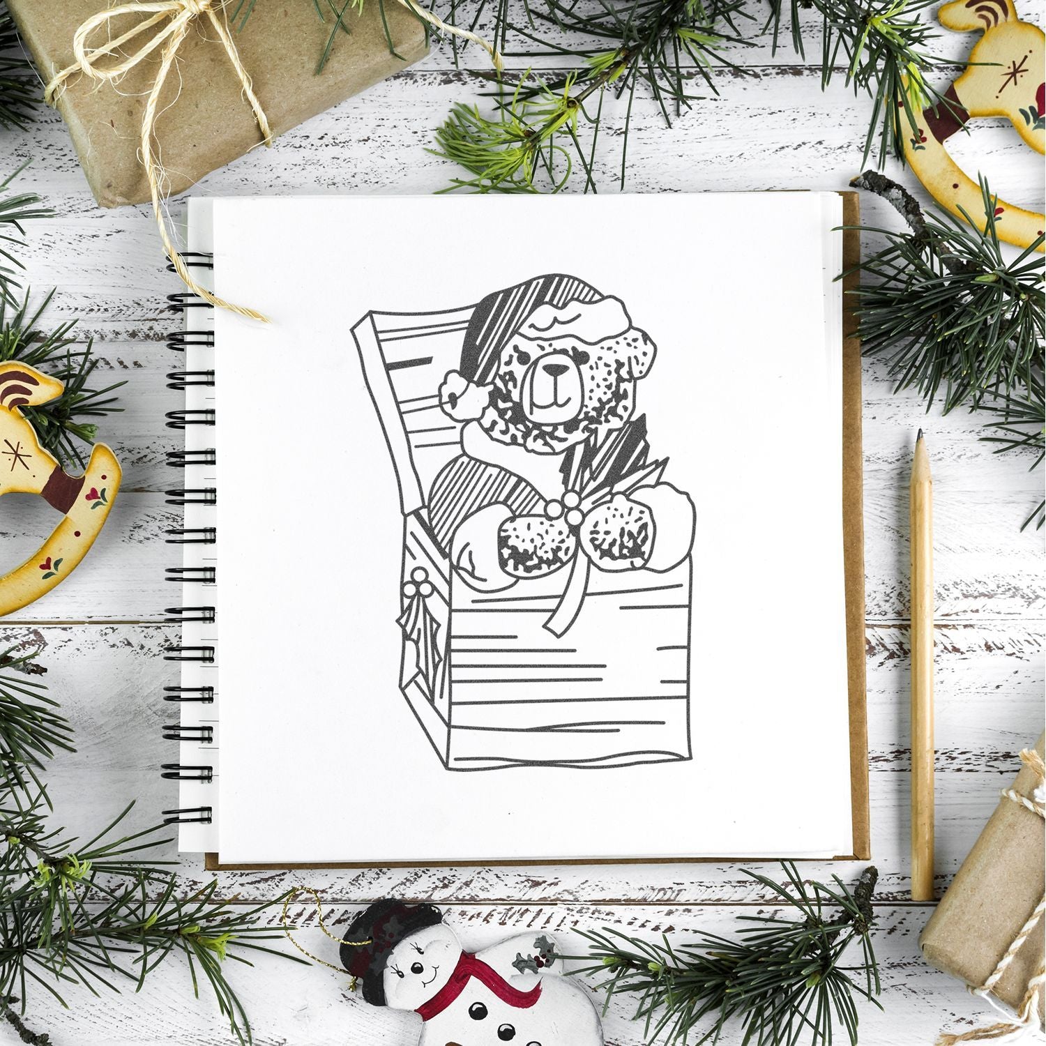 Christmas-themed sketchbook with teddy bear Santa drawing, surrounded by pine branches, gift wrapping, and holiday ornaments on a rustic white table.