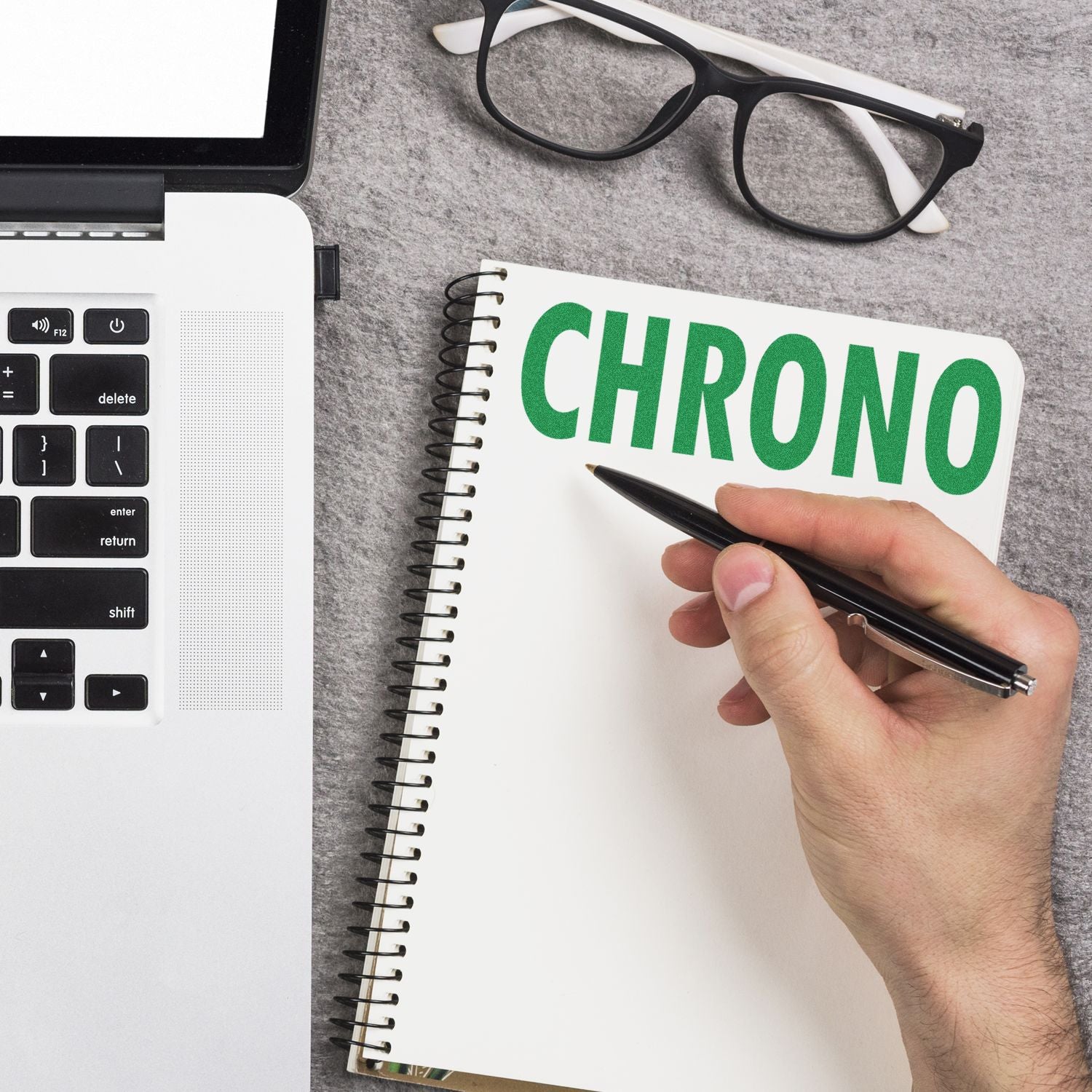 Hand writing CHRONO in a notebook beside a laptop and eyeglasses, concept of time management and productivity.