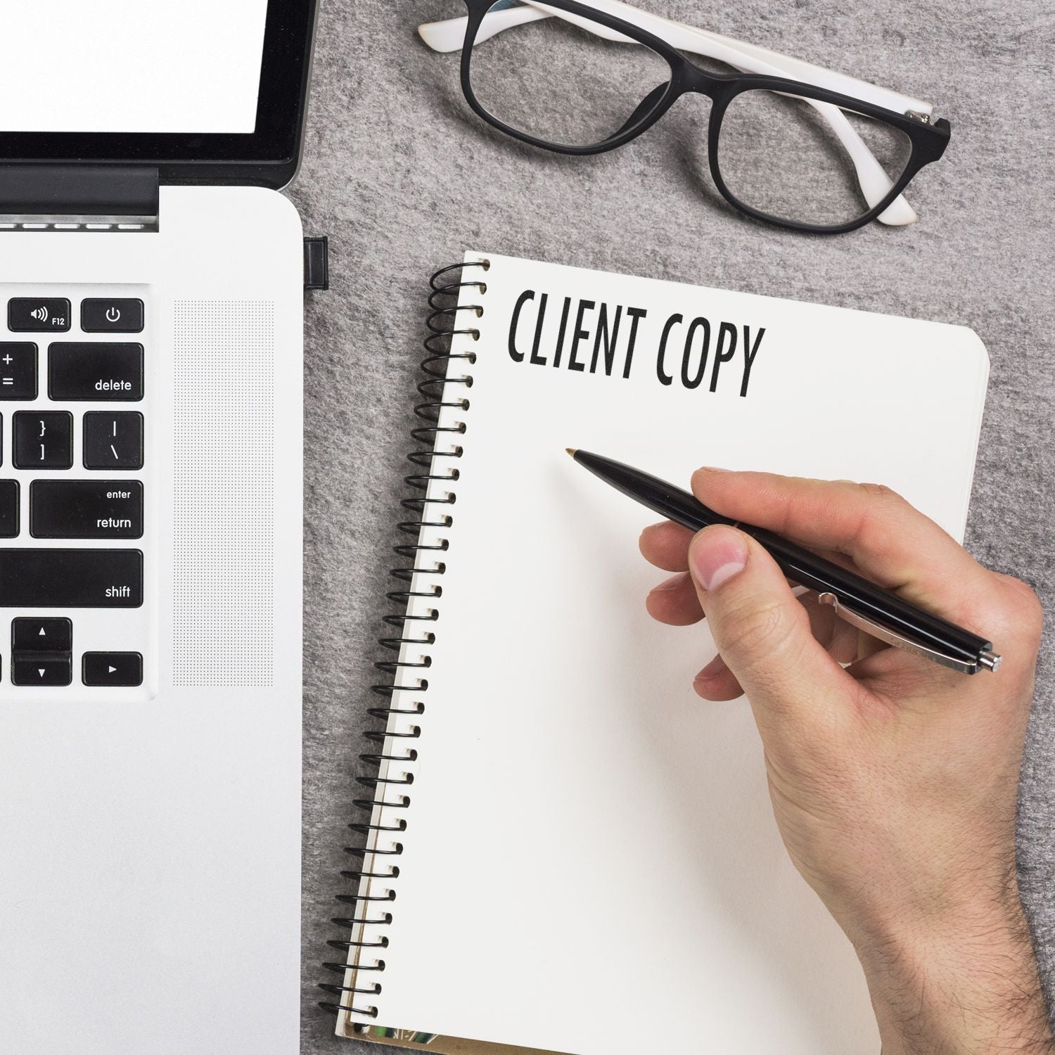 Hand writing in a client copy notebook beside a laptop and eyeglasses on a modern office desk.