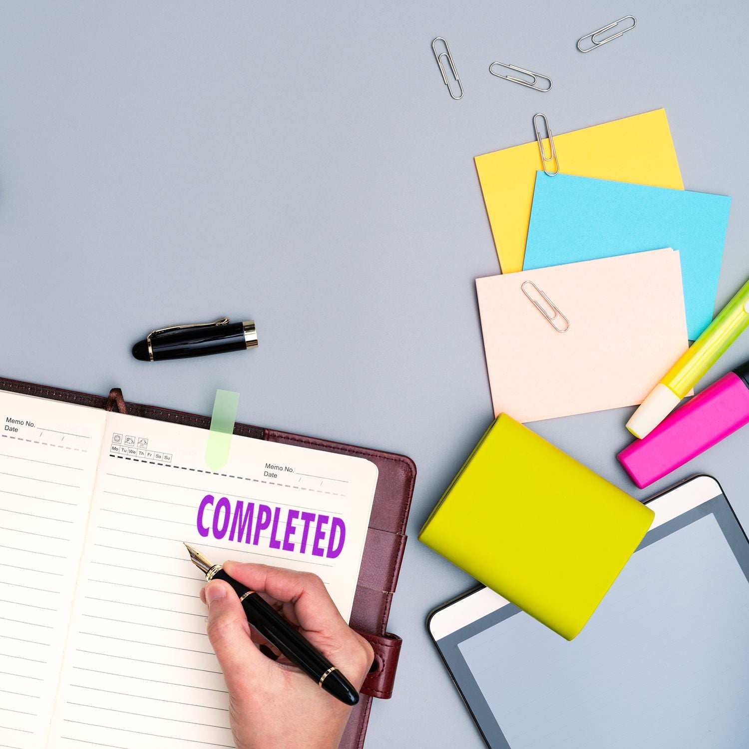 Hand writing in planner with COMPLETED marking task success on desk with stationery, notes, and colorful memo folders for productivity