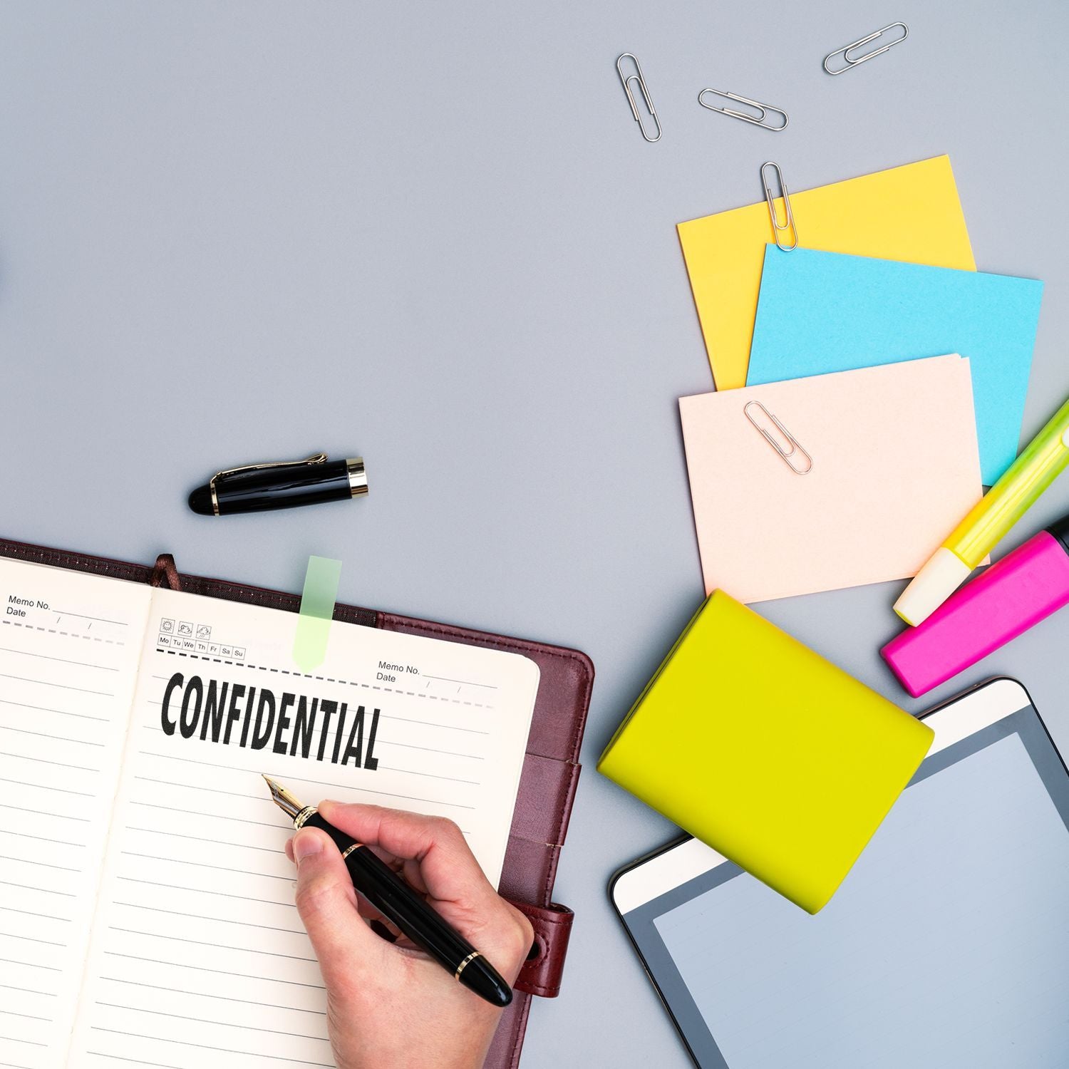 Hand writing confidential in a notebook with colorful sticky notes, pen, paper clips, and office stationery on a desk.