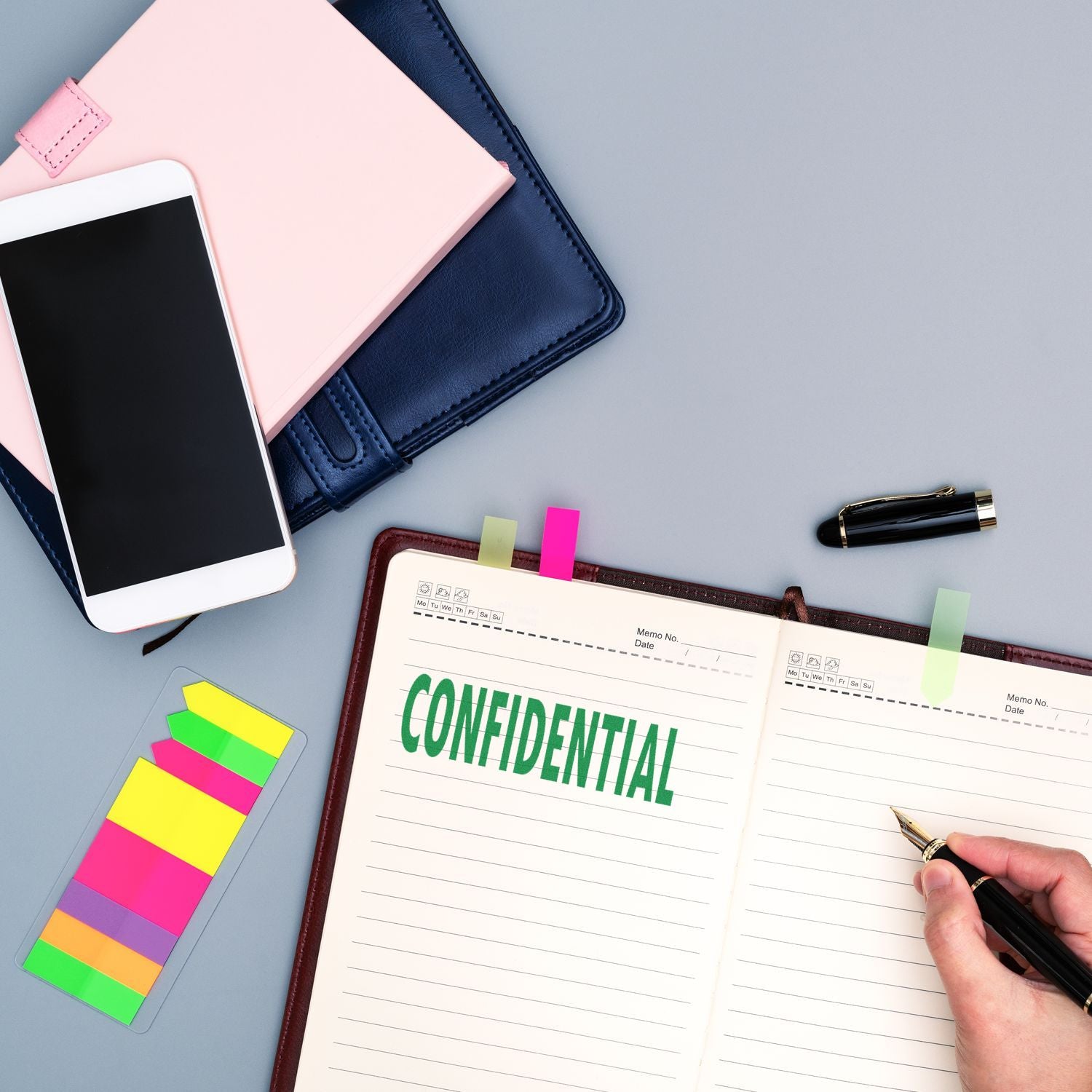 Top view of office desk with smartphone, notebooks, colorful sticky notes, and an open confidential planner being written in with a pen.