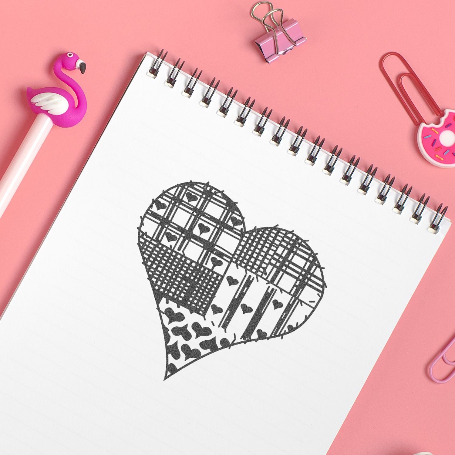 Cute pink stationery flat lay with spiral notebook featuring a patterned heart drawing, flamingo pen, binder clips, and paper clips.