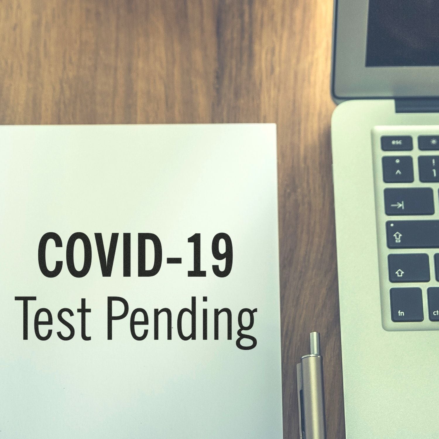 COVID-19 test pending notice on paper beside laptop keyboard and pen, indicating awaiting results in an office.
