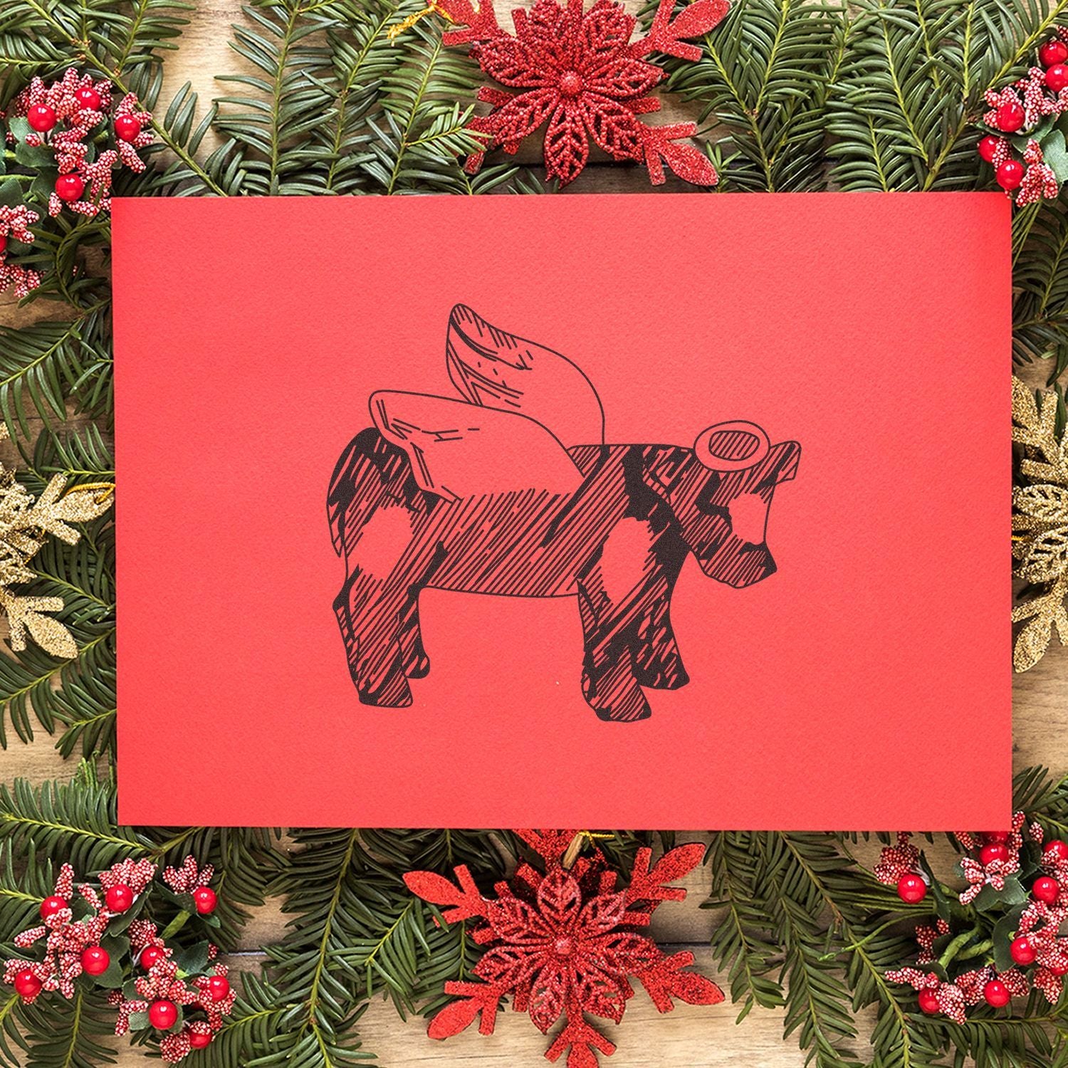 Red Christmas greeting card with a winged cow illustration, framed by festive evergreen garland, berries, and snowflake ornaments on a rustic wood background.