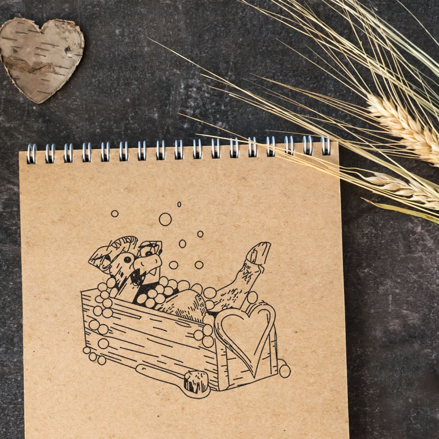 Rustic spiral notebook with sketch of bread and a heart-shaped wooden box on a dark background with wheat stalks.