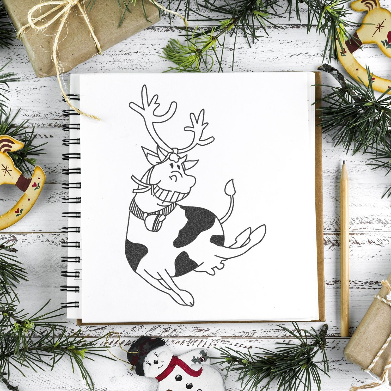 Christmas cow reindeer sketch in notebook with festive holiday decor, pine branches, and wrapped gifts on rustic white wood background.