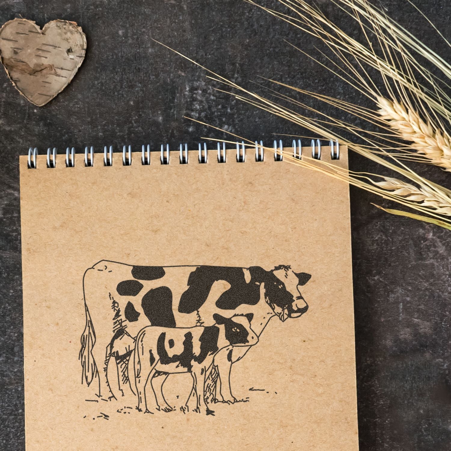 Rustic kraft notebook with cow and calf sketch, wheat stalks, and a birch heart on a dark textured background.