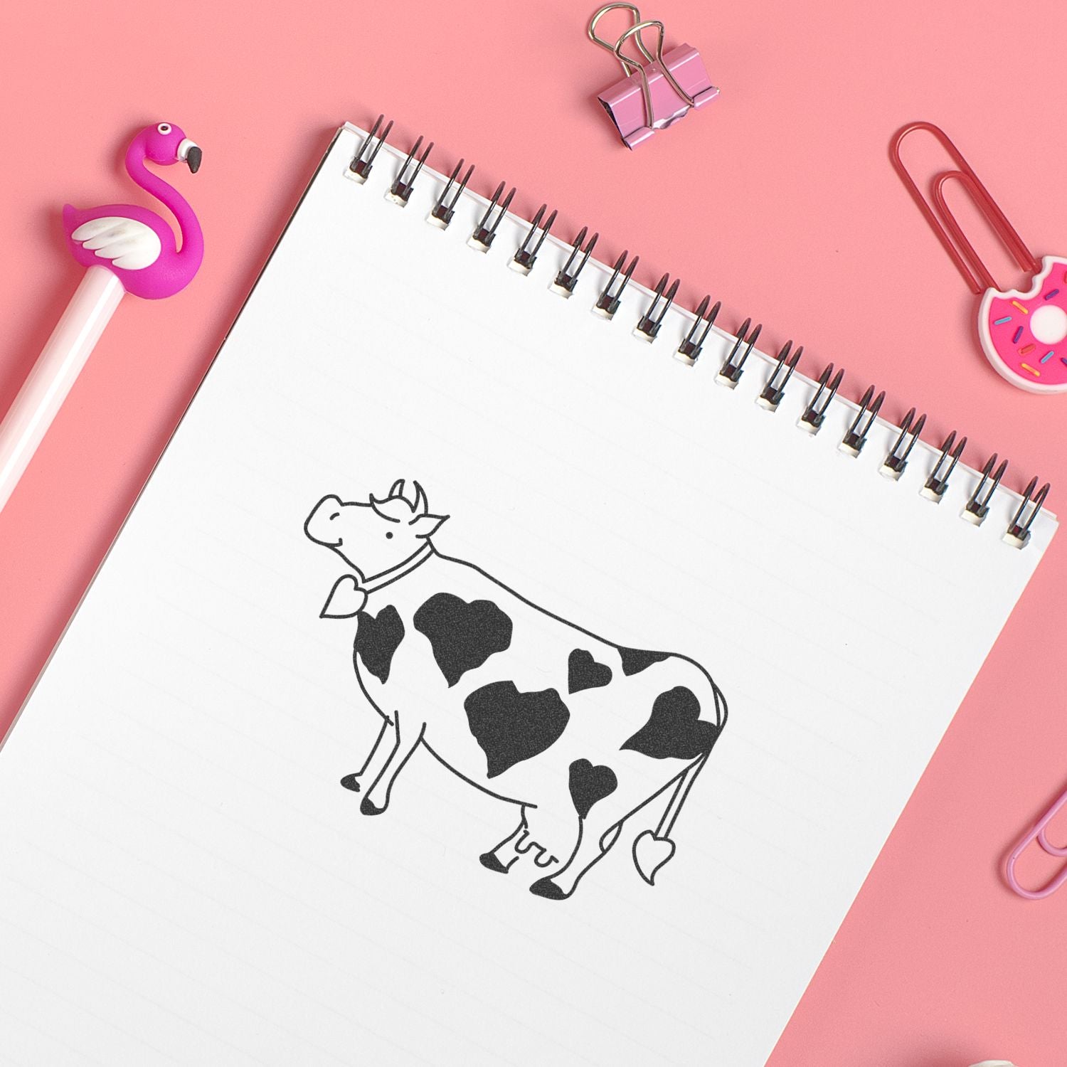 Cute spiral notebook with cow doodle on pink background, surrounded by flamingo and donut paper clips and stationery accessories.