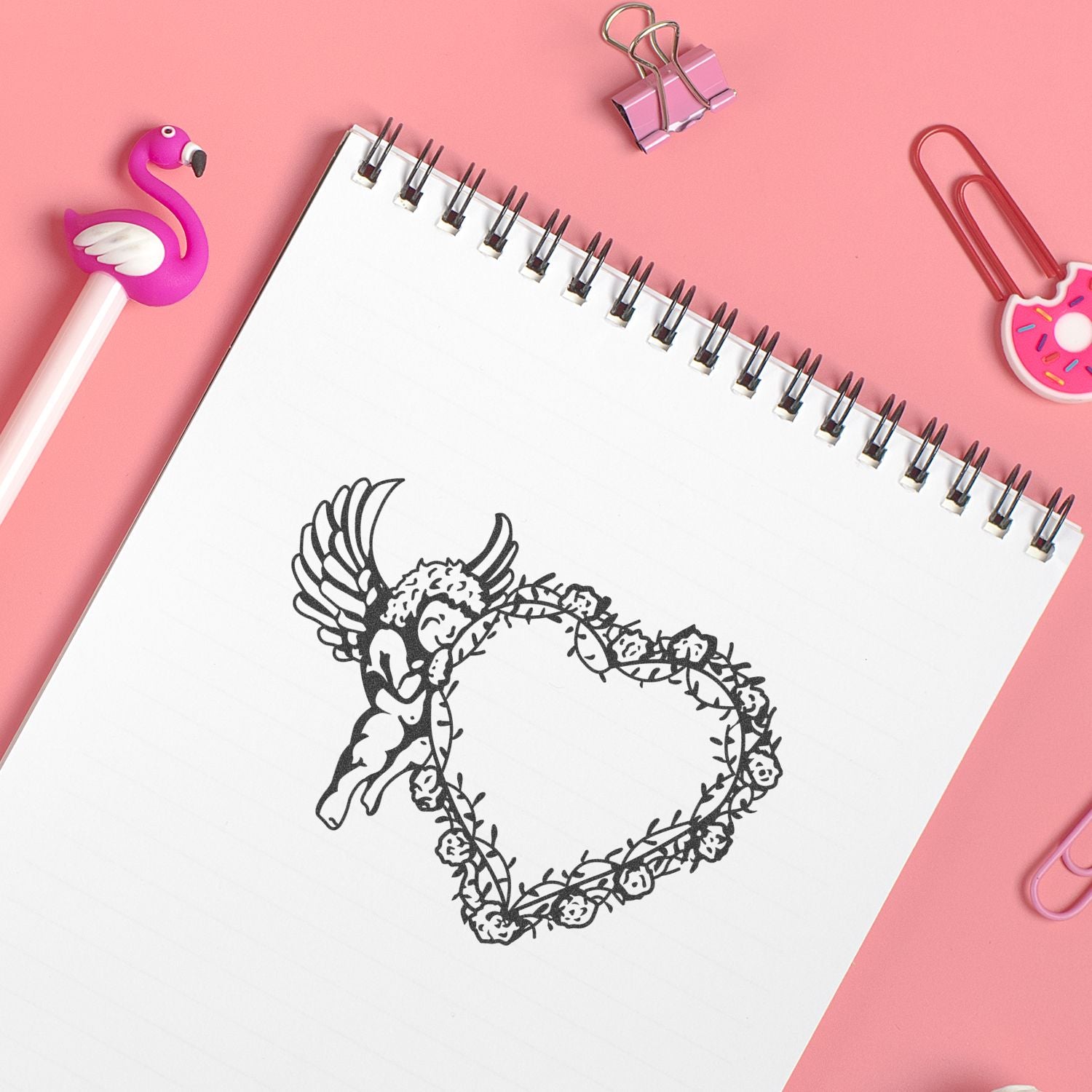 Cute stationery flat lay with spiral notebook featuring an angel heart design on a pink background, with flamingo pen and decorative paper clips.