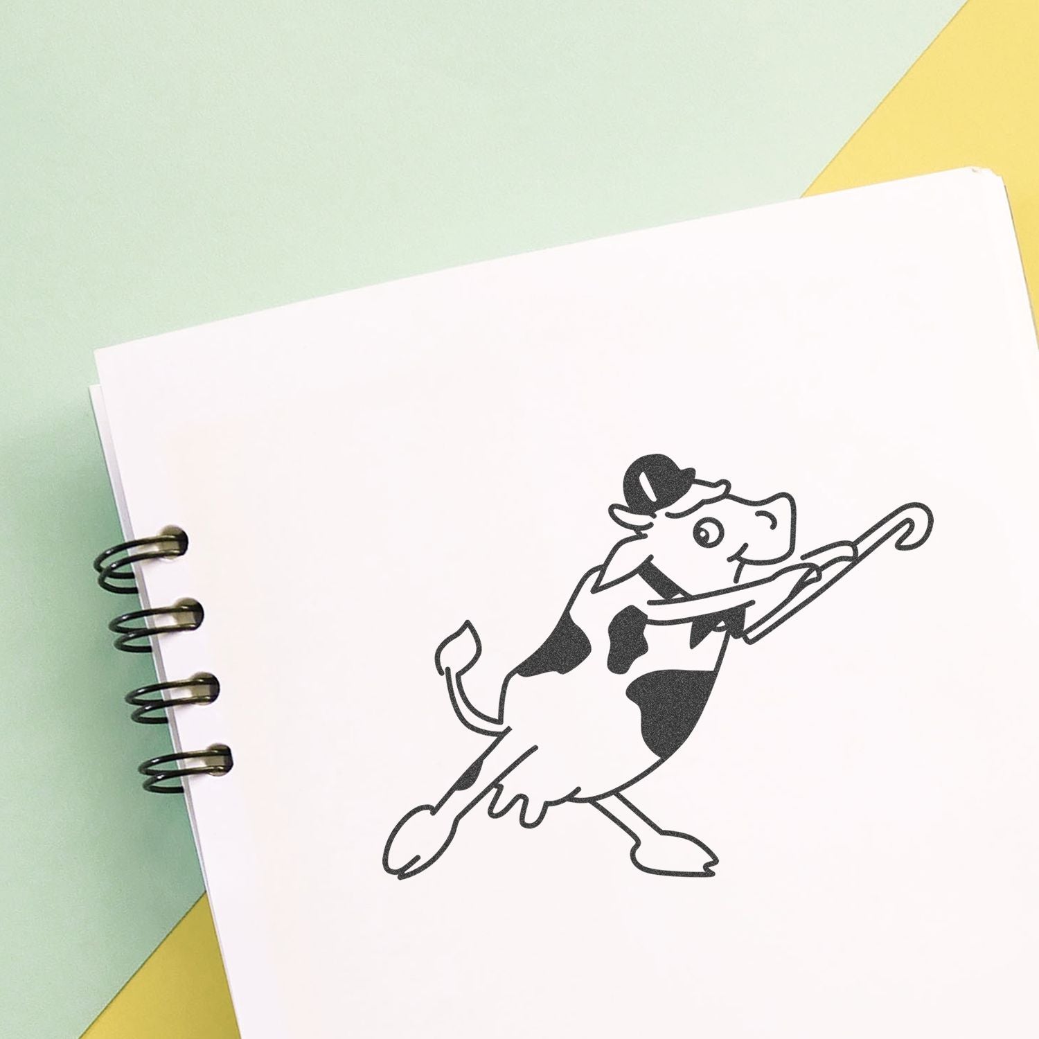 Minimalist spiral notebook with a playful cow logo illustration on a pastel green and yellow background.