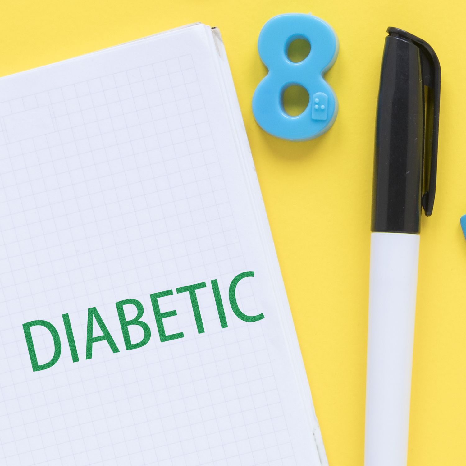 Diabetes awareness notebook with DIABETIC lettering, pen, and blue 8 eraserhealth care reminder on a yellow background.
