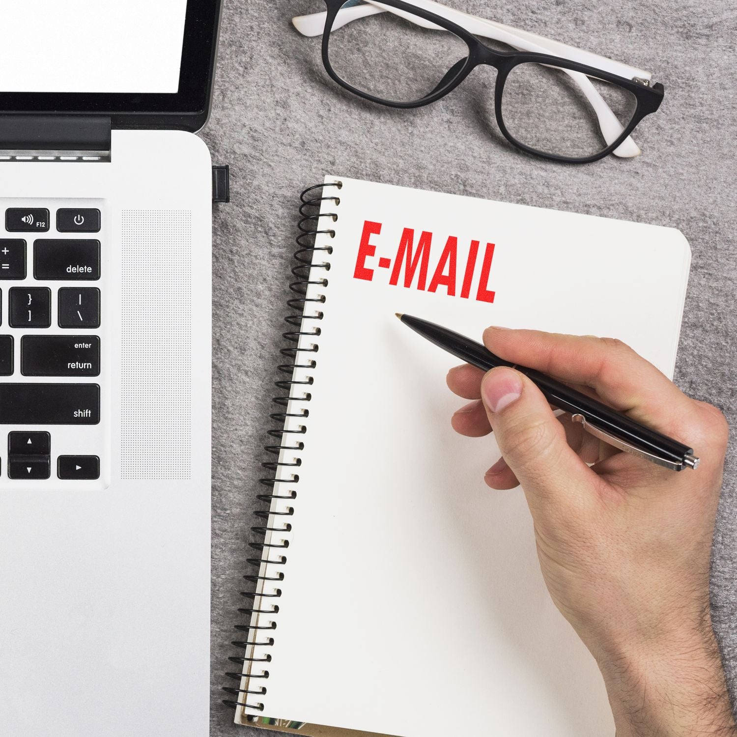 Hand writing e-mail in notebook beside laptop and eyeglasses on office desk.