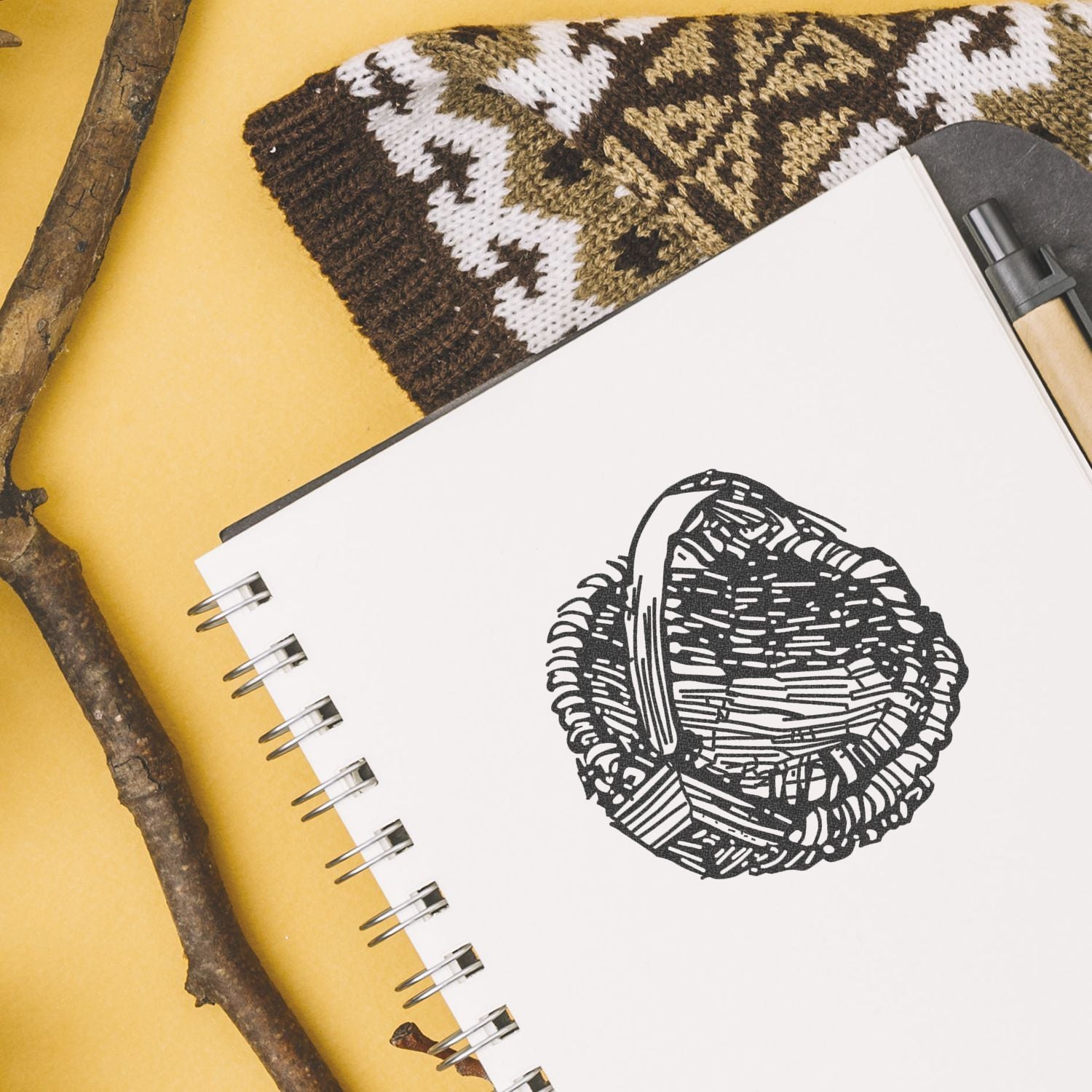 Spiral notebook with a black woven basket illustration on a yellow background, styled with rustic sticks and knitted textile.