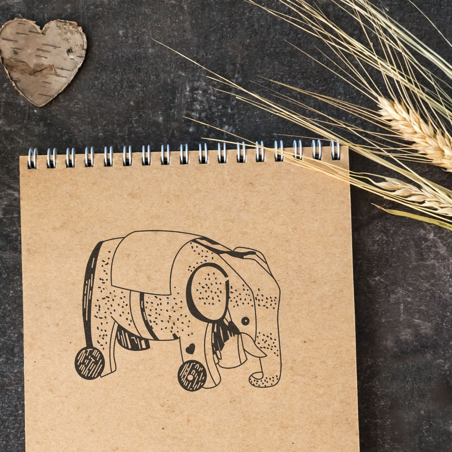 Eco-friendly spiral notebook with elephant illustration on rustic dark background, styled with dried wheat and birch heart decor.